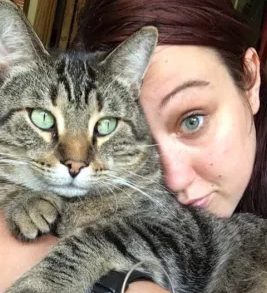 A woman with light skin and red hair holds a large tabby cat with green eyes close to her face.
