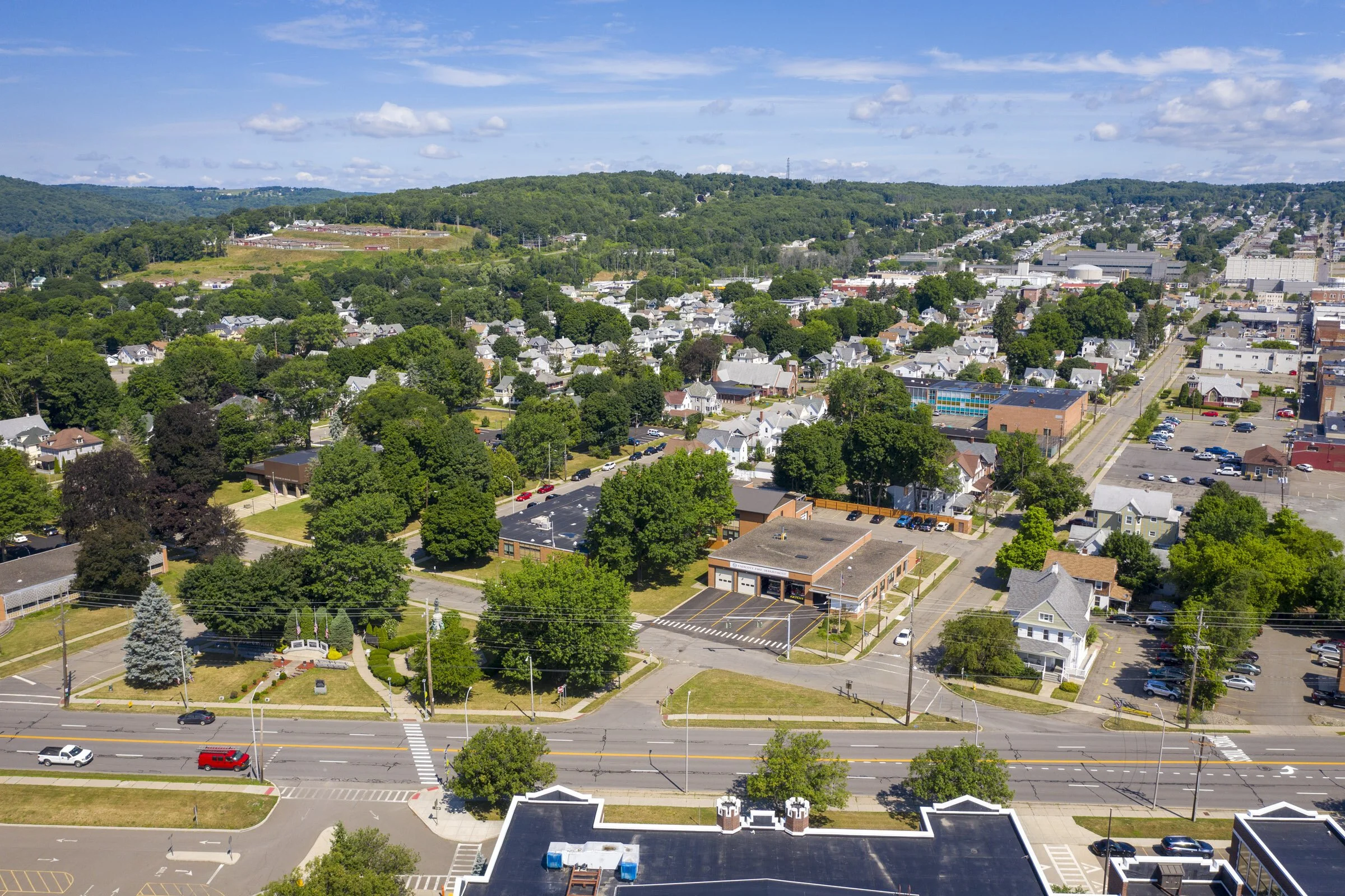 Aerial view of a town with residential houses, commercial buildings, trees, and roads, surrounded by green hills and a blue sky with scattered clouds.