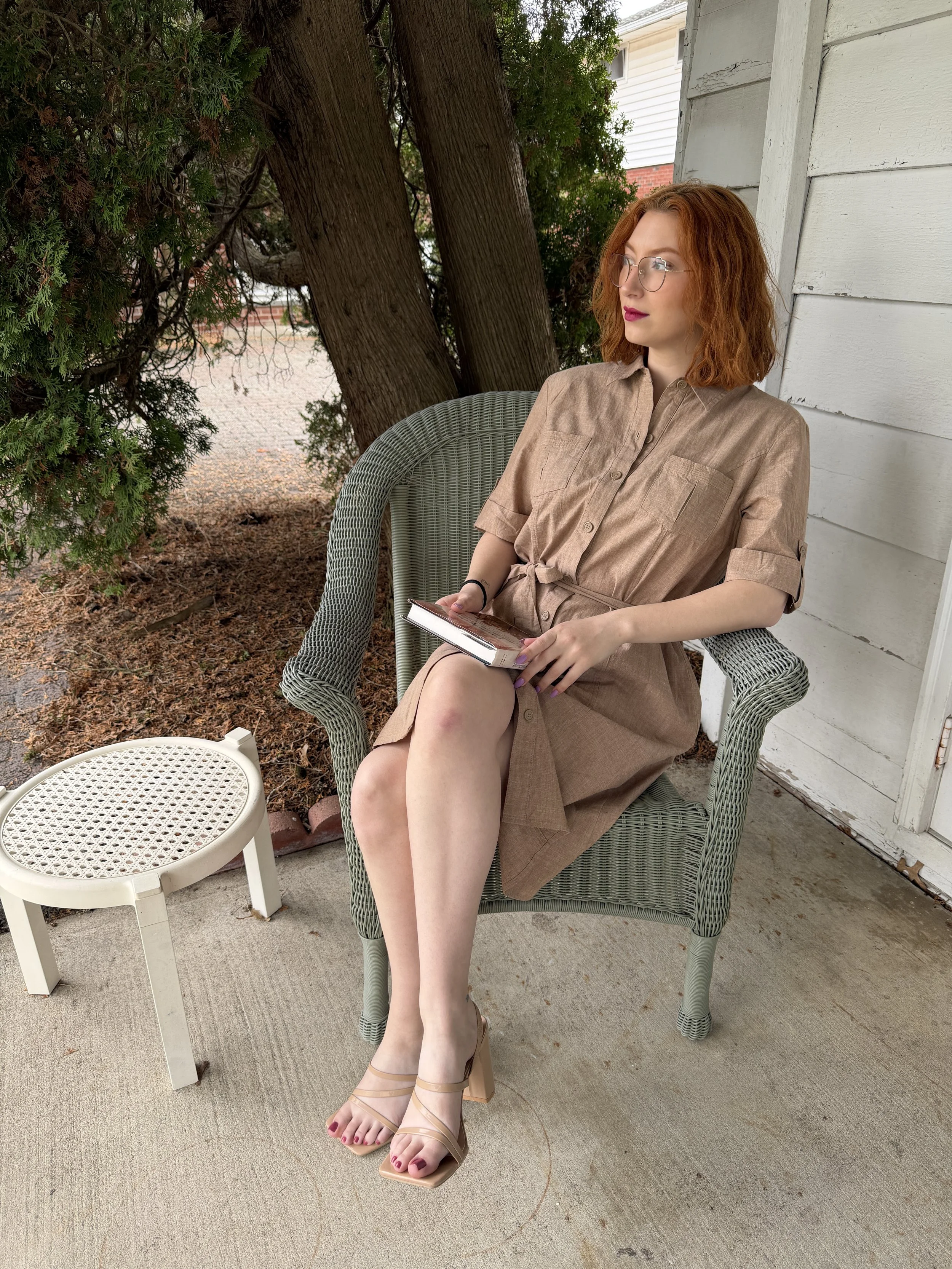 A young woman with red curly hair, glasses, wearing a tan dress, sitting outside on a patio chair, holding a book, with trees and a white house in the background.
