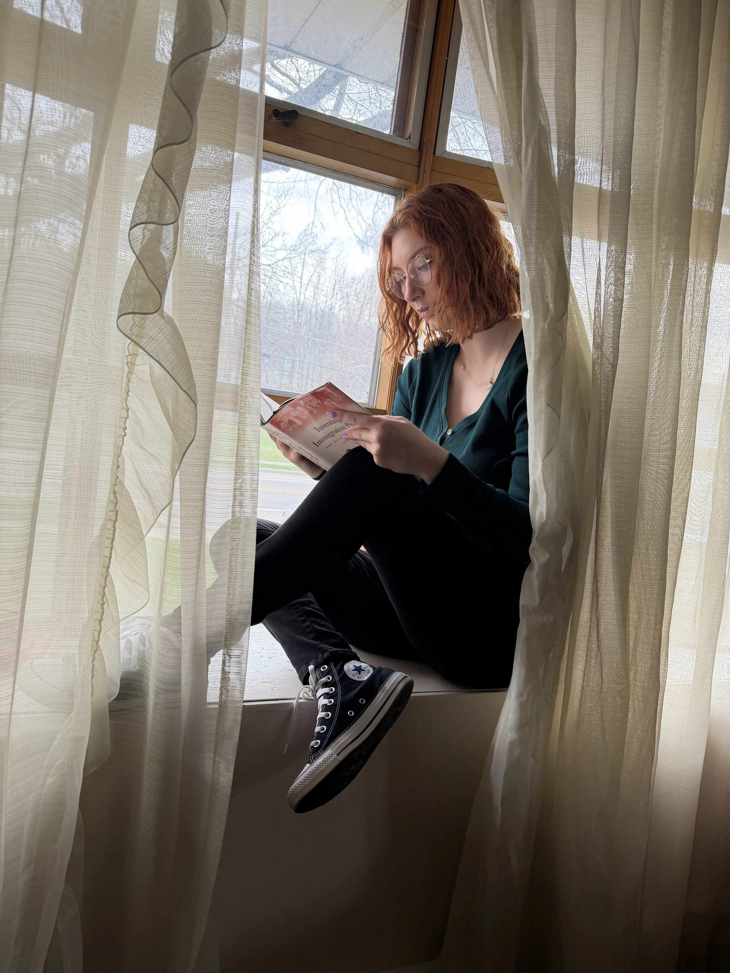 A young woman with curly red hair and glasses sitting on a windowsill, reading a book, with sheer curtains partially covering her, and trees visible outside through the window.