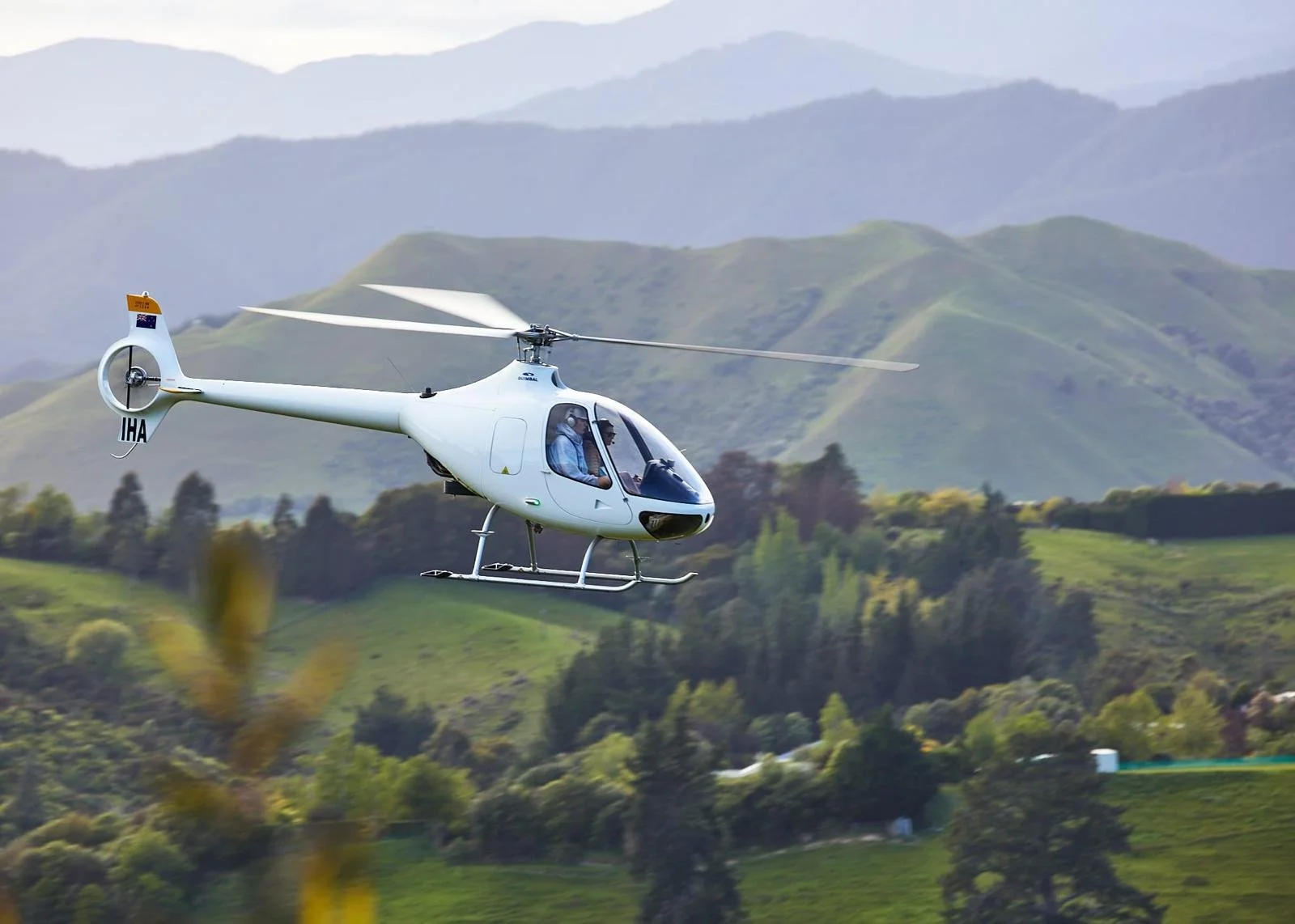 A man standing next to a white helicopter on a riverbank with a forested mountain in the background.