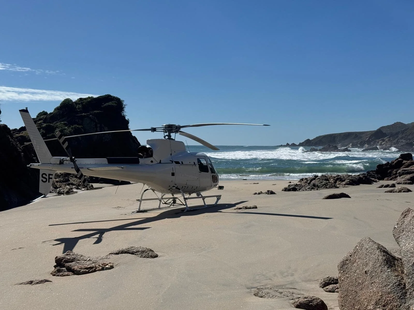 A helicopter on a sandy beach near rocks with the ocean and cliffs in the background.