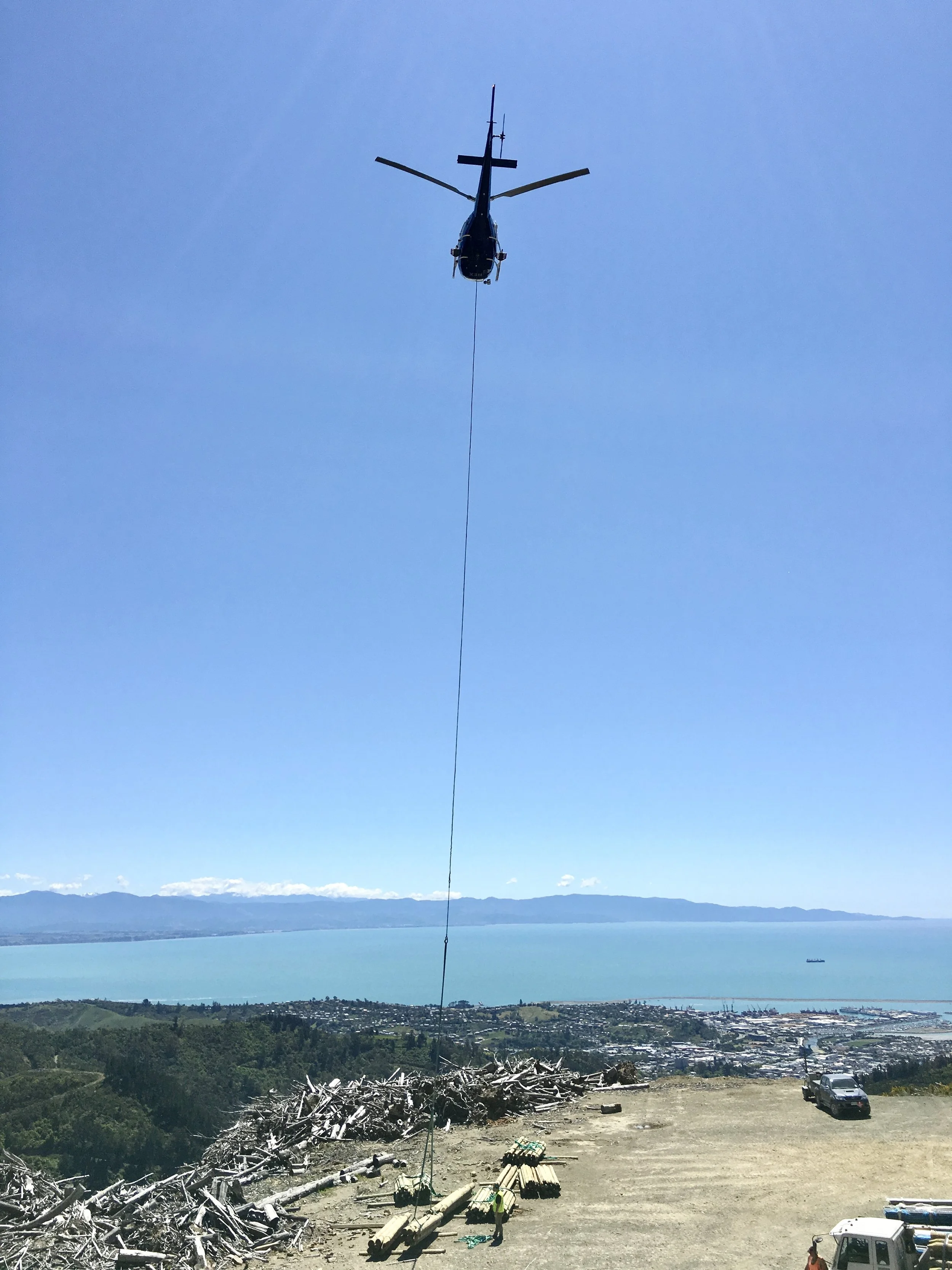 A helicopter flying in the clear blue sky over a landscape, with a long cable hanging from it, possibly for delivering or lifting objects.