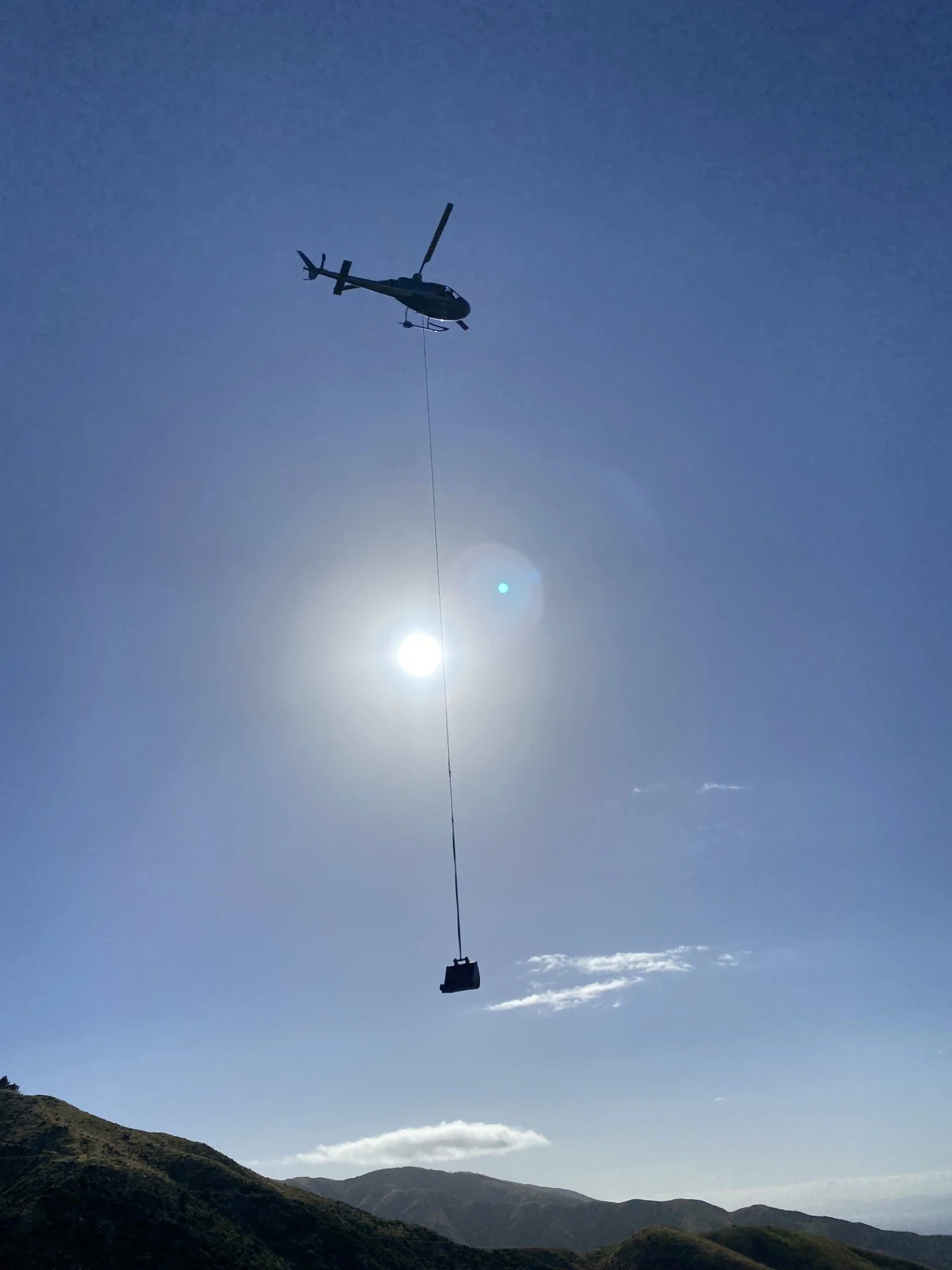 Helicopter flying in the sky with a cable and weight hanging from it, over a mountain landscape with the sun behind it.