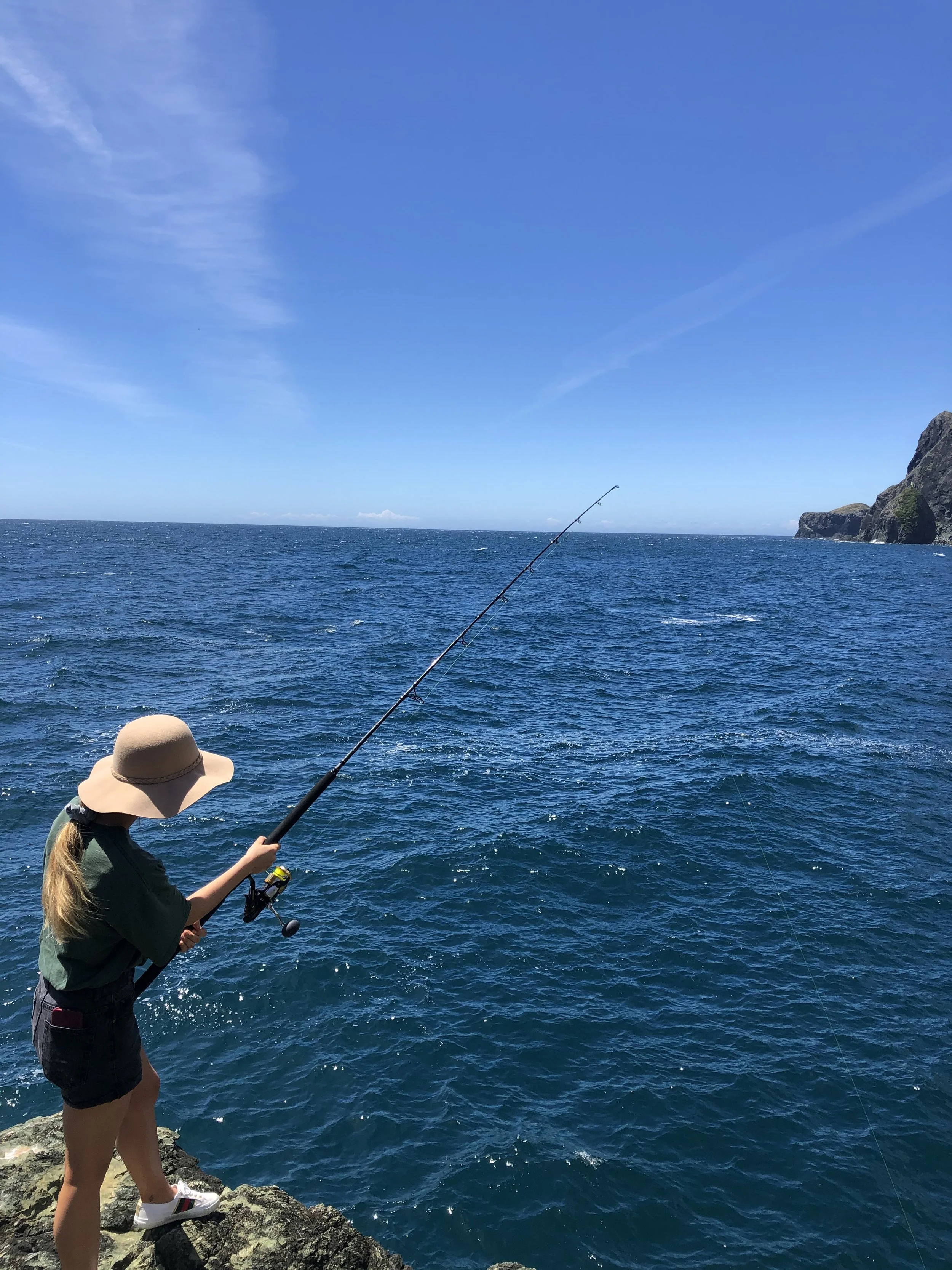 A young girl wearing a wide-brimmed hat, green jacket, black shorts, and sneakers standing on rocks near the ocean, fishing with a rod on a bright, sunny day. The ocean is blue with gentle waves, and there are cliffs in the distance under a partly cloudy sky.