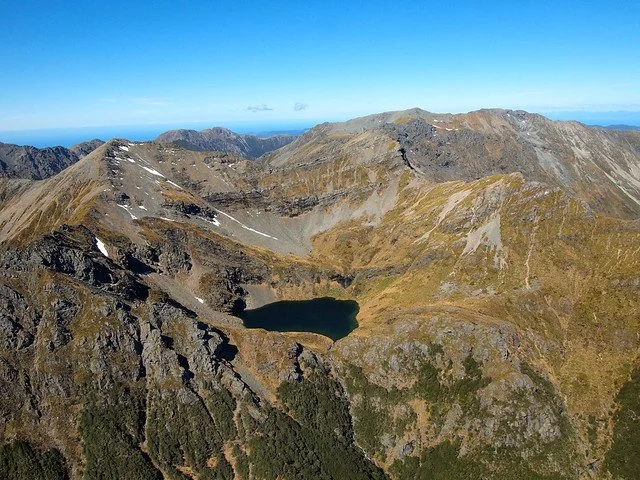A scenic view of a lake with mountains in the background, a dock extending into the water, trees along the shoreline, and a helicopter flying in the clear blue sky.