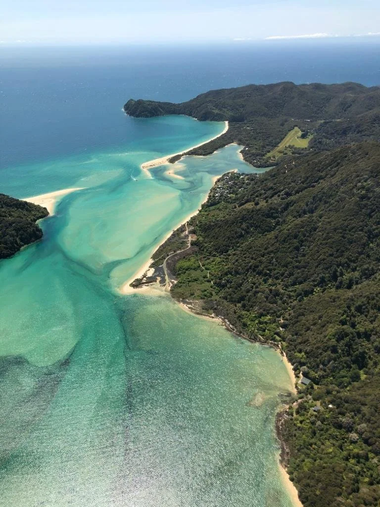 Aerial view of a coastal bay with sandy beaches, clear turquoise water, green hills, and dense forested land.
