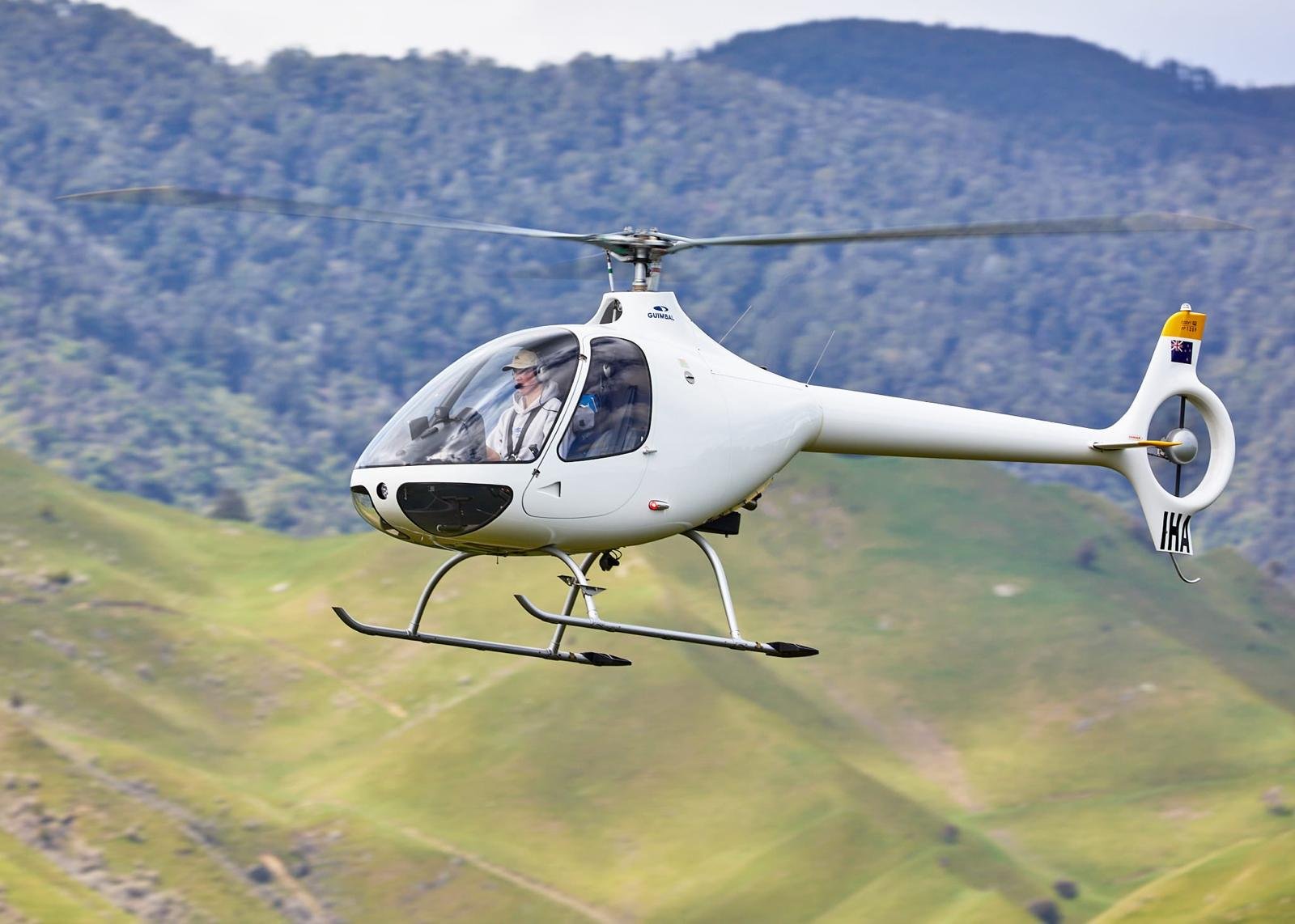 A white helicopter flying over green grassy hills with a mountainous background.