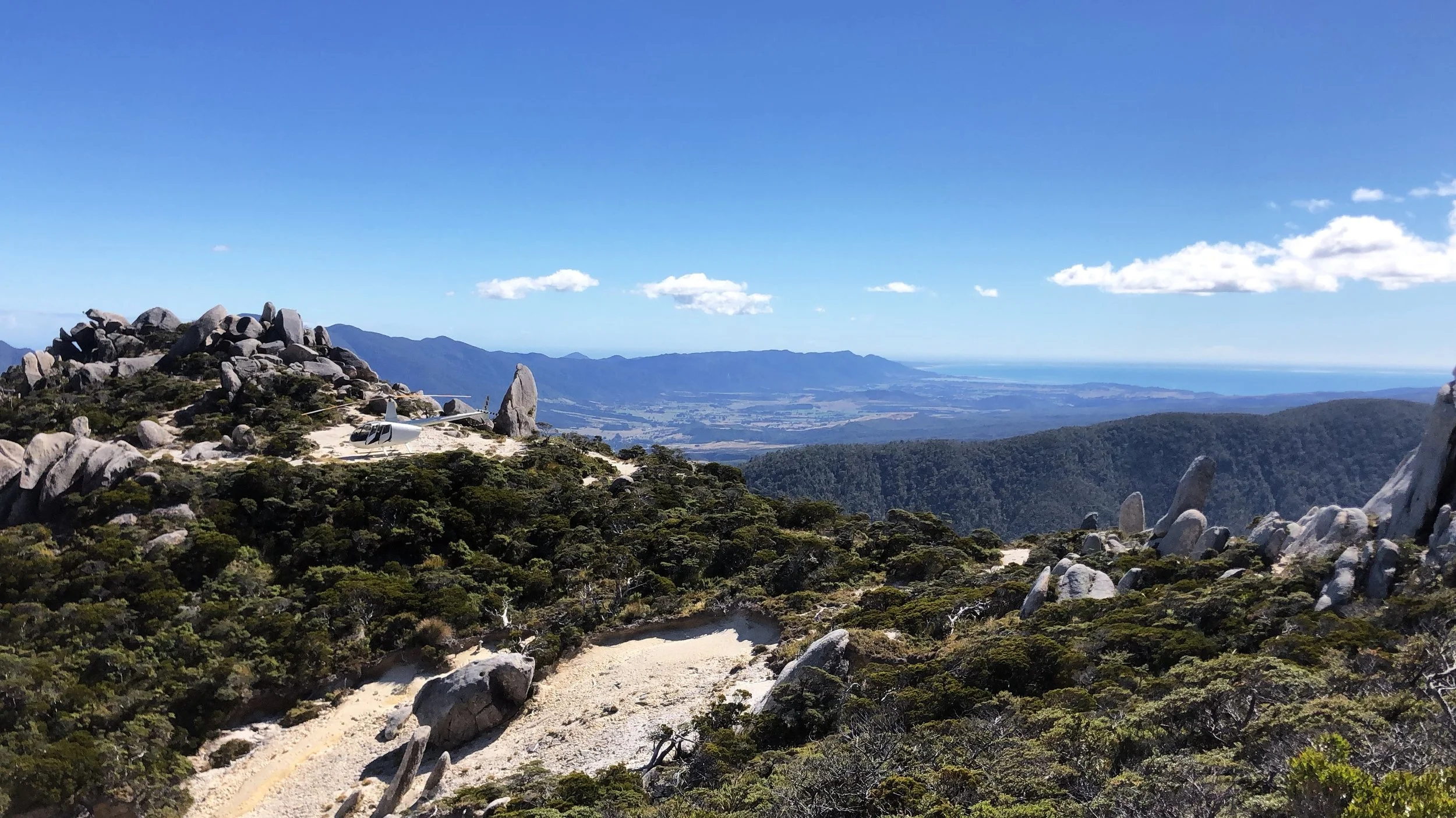 Scenic mountain landscape with rocky terrain, a helicopter on a dirt trail, lush green bushes, and distant mountains under a blue sky with scattered clouds.