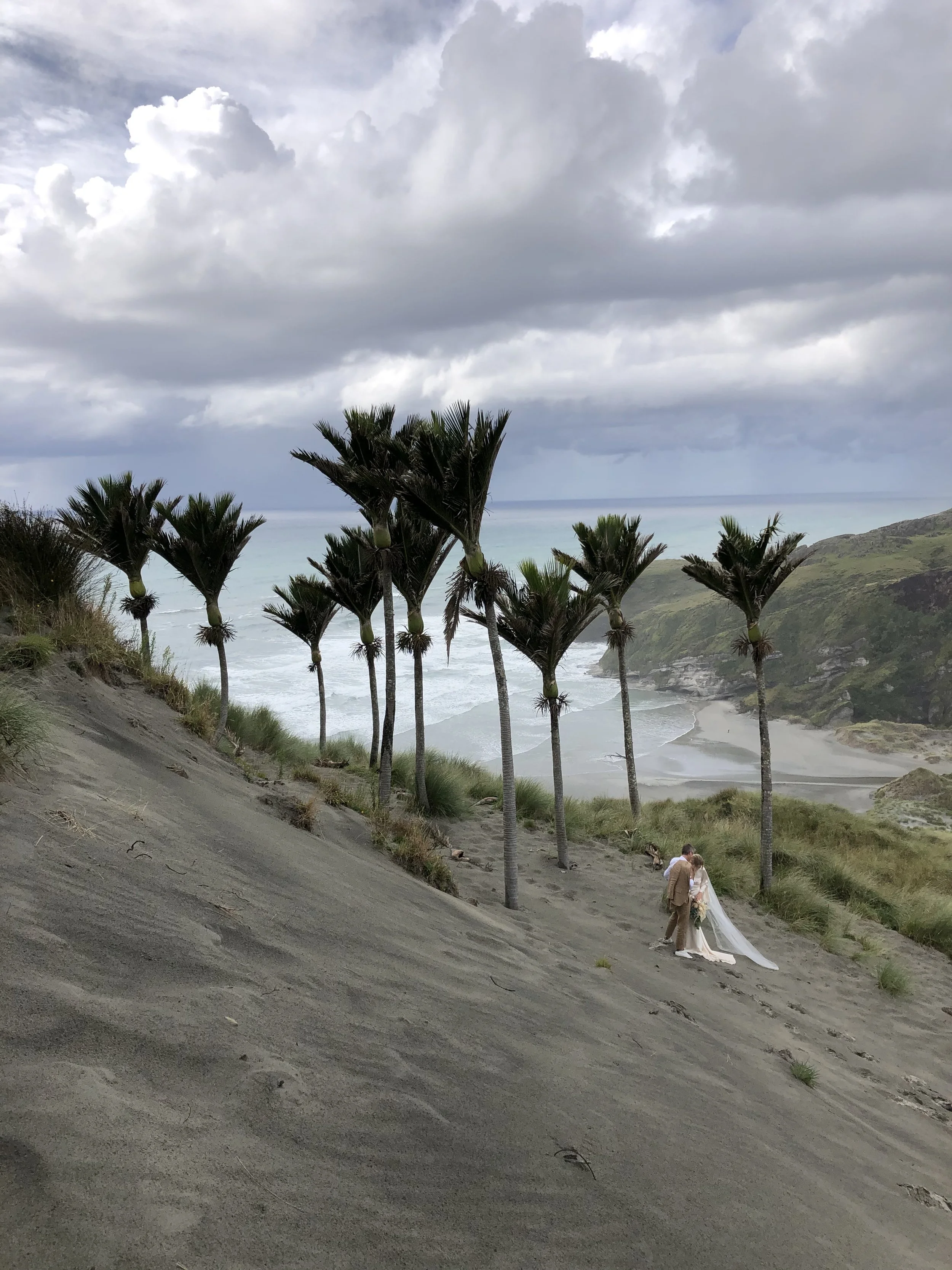 A black helicopter on a beach next to a large cliff. In the background, a couple is dancing with a photographer taking pictures, with the ocean and cloudy sky behind them.