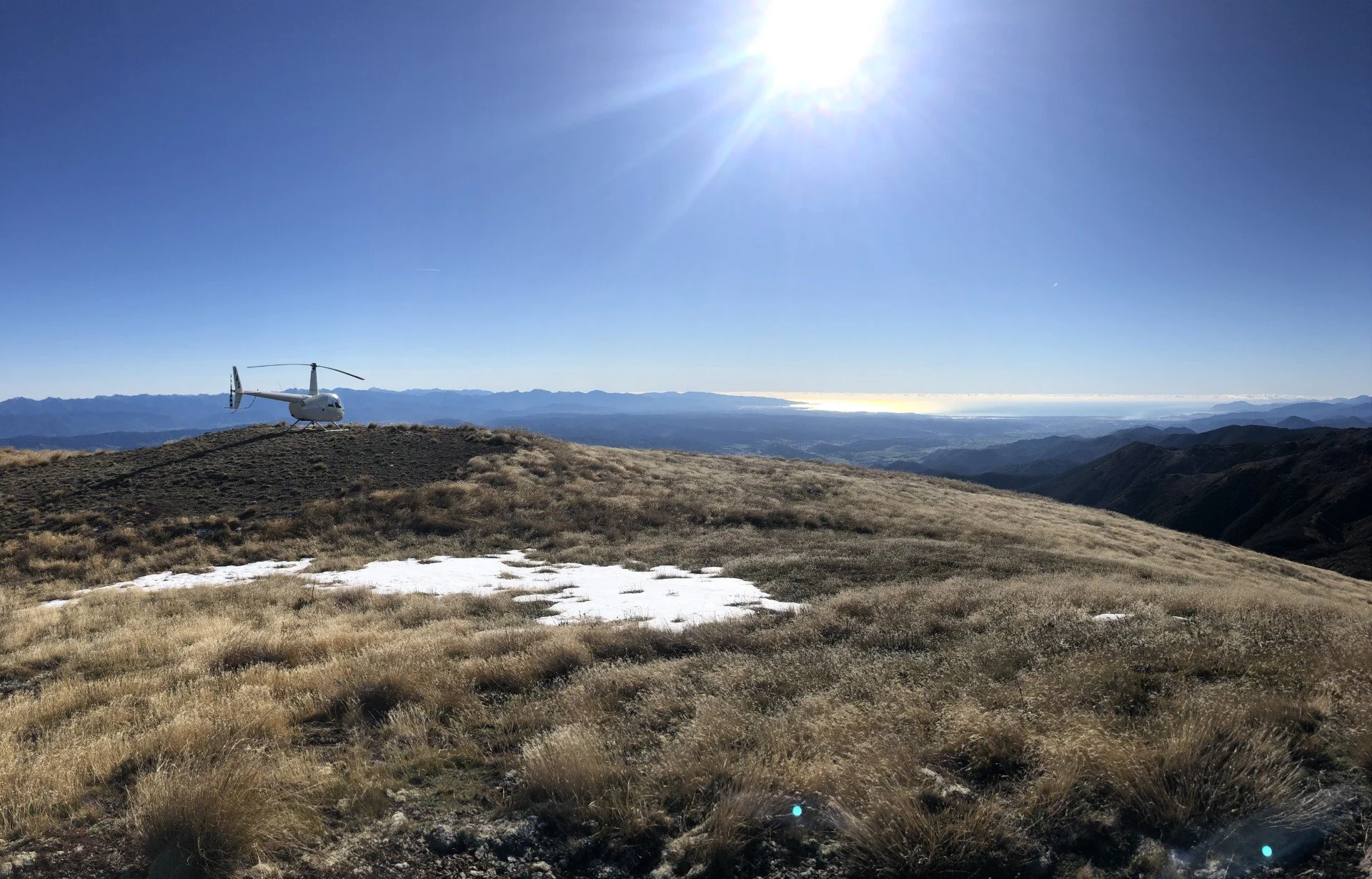 A helicopter parked on a grassy hilltop with mountains and a body of water in the distance under a clear blue sky and bright sun.