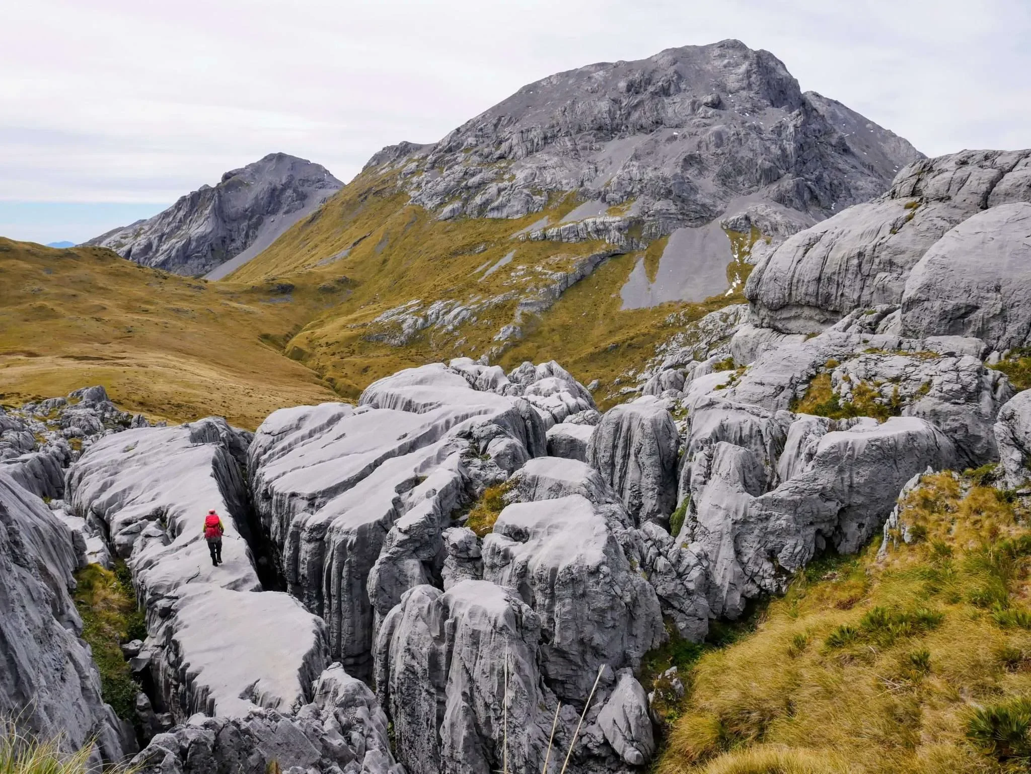 A hiker in a red jacket walking through a narrow rocky gorge with large gray rocks, with mountains in the background and a cloudy sky above.