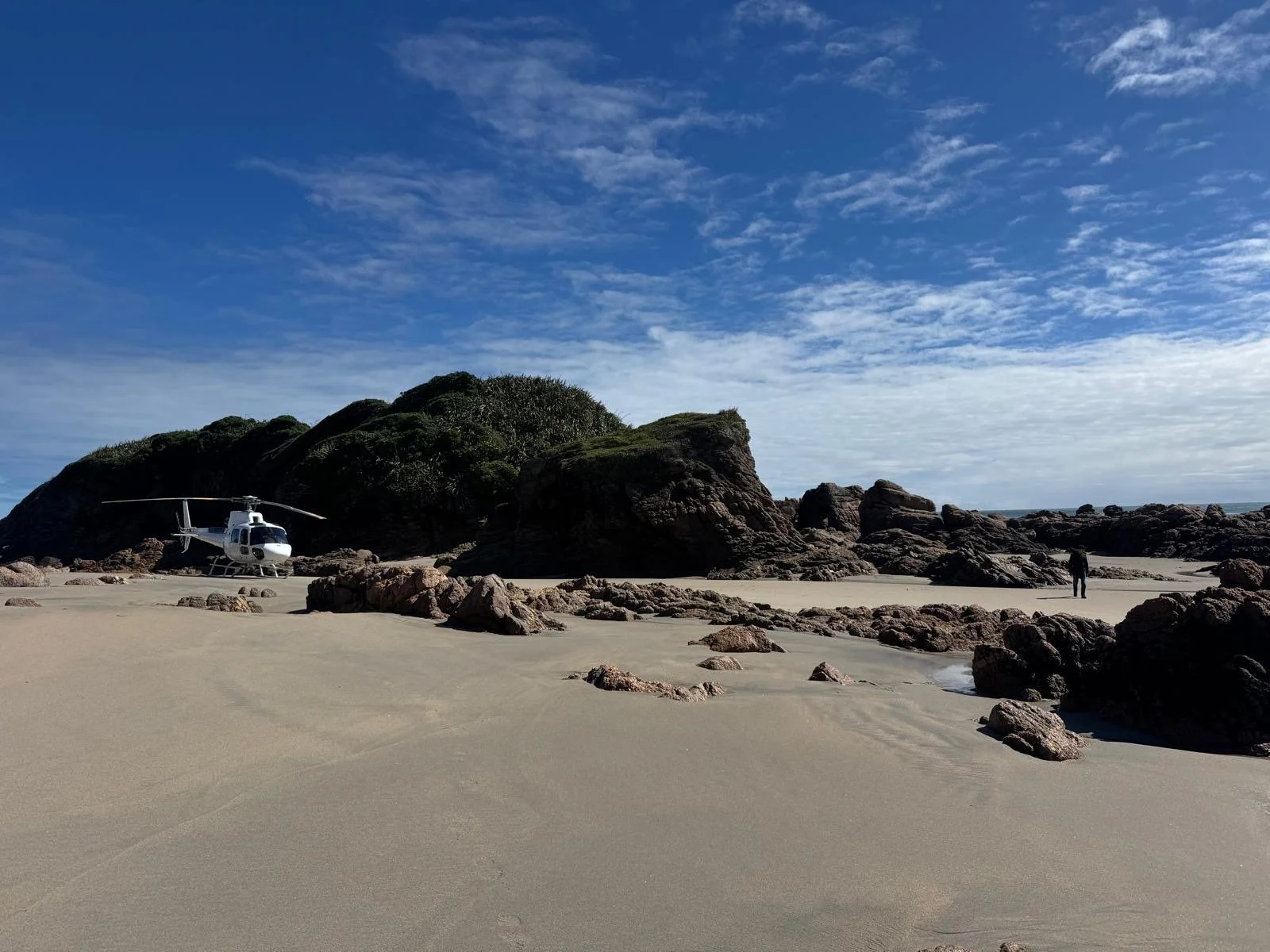 A helicopter parked on a sandy beach near a rocky outcrop with a person walking in the distance under a partly cloudy sky.