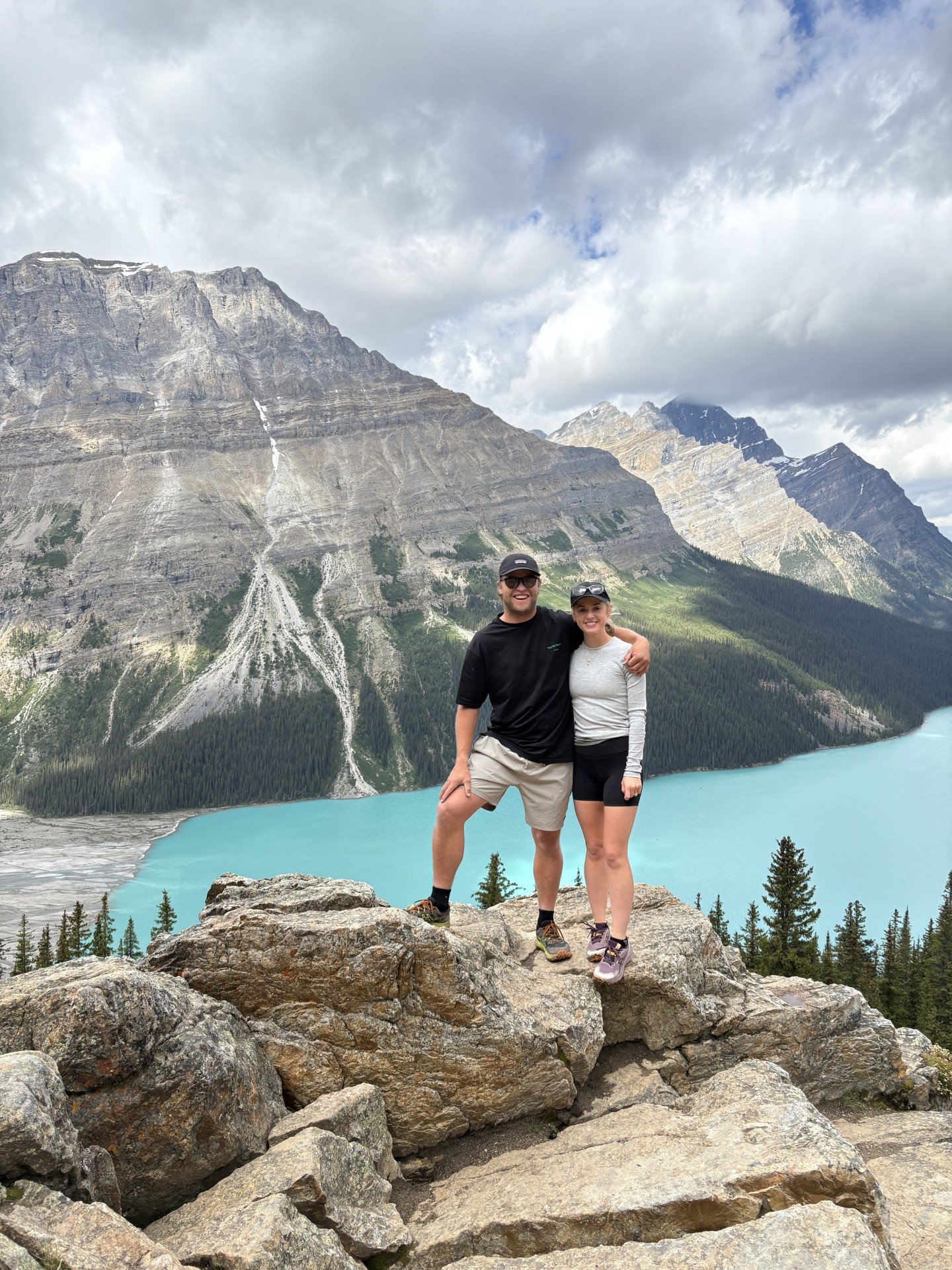A smiling man and woman standing together on a rocky ledge in front of a turquoise lake, with steep mountain peaks and partly cloudy sky in the background.