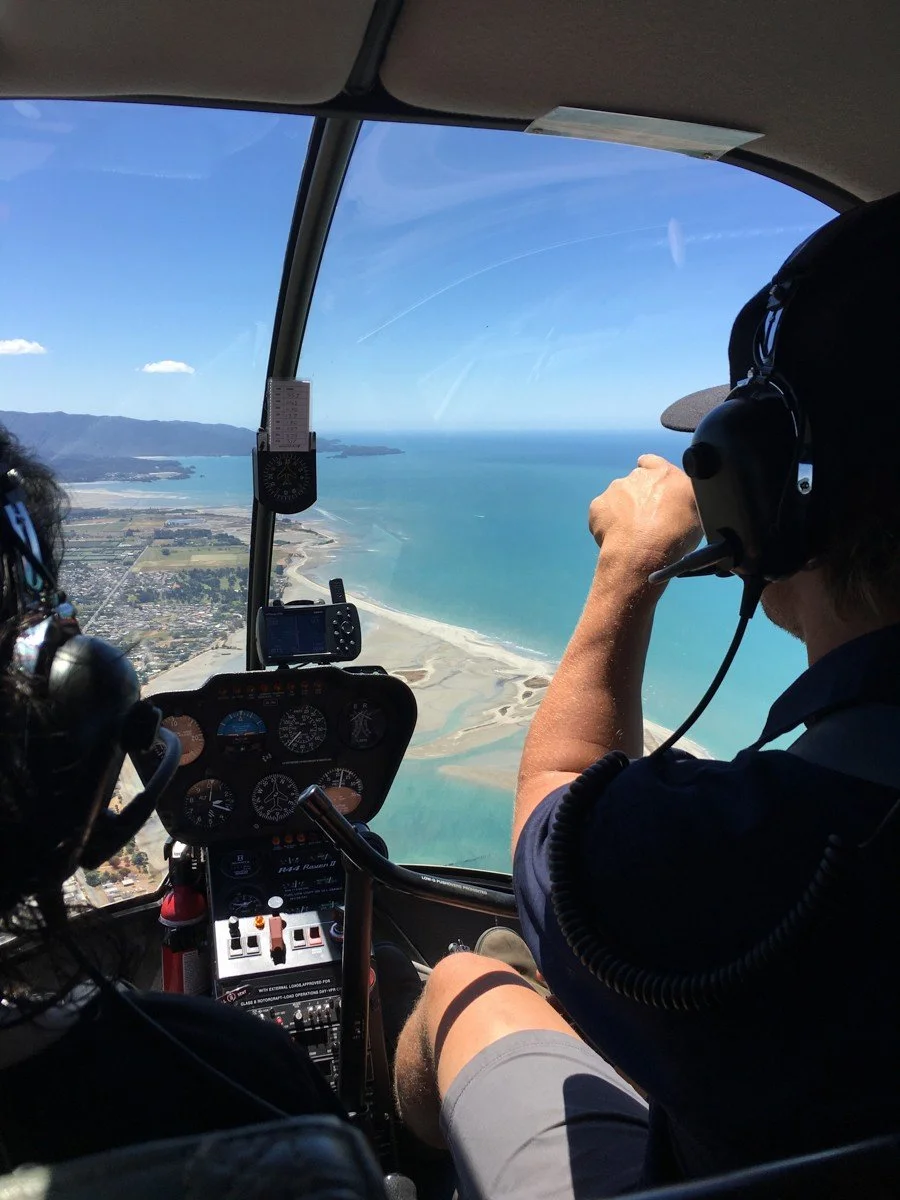 View from inside a helicopter cockpit showing two pilots flying over a coastal landscape with a beach and water.
