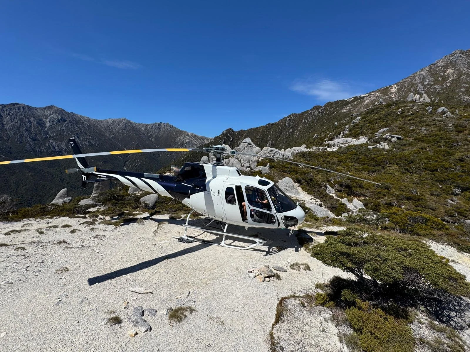 A black and white helicopter on rocky terrain in a mountainous area with clear blue sky and sparse green bushes.
