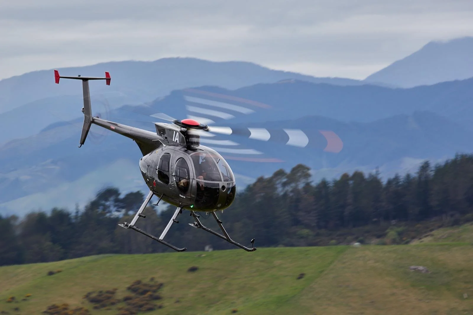 Helicopter flying in the sky with a cable and weight hanging from it, over a mountain landscape with the sun behind it.