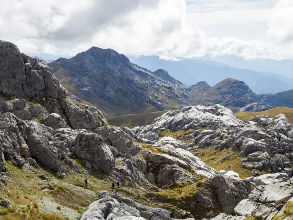 A mountain landscape with rocky terrain, a helicopter on a trail, lush green bushes, and a distant view of mountains and coastline under a blue sky with scattered clouds.
