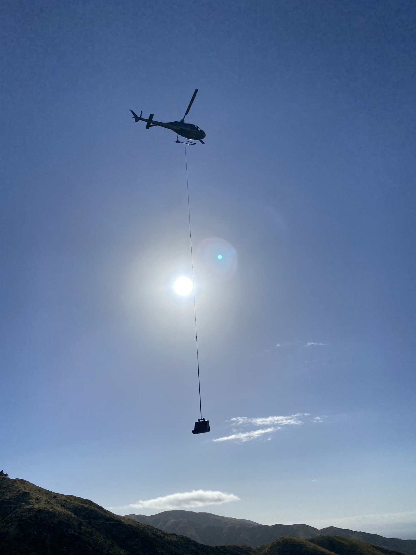 Utility helicopter flying in the clear blue sky, towing a large object on a cable over hilly landscape.
