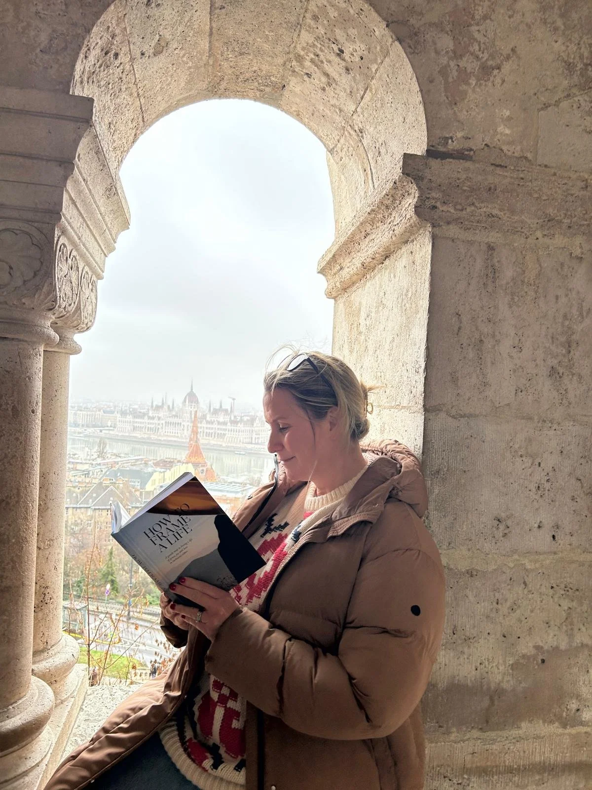 A woman reading a book near a stone archway with a view of Budapest, including the Parliament building and the Eiffel Tower replica, in the background.