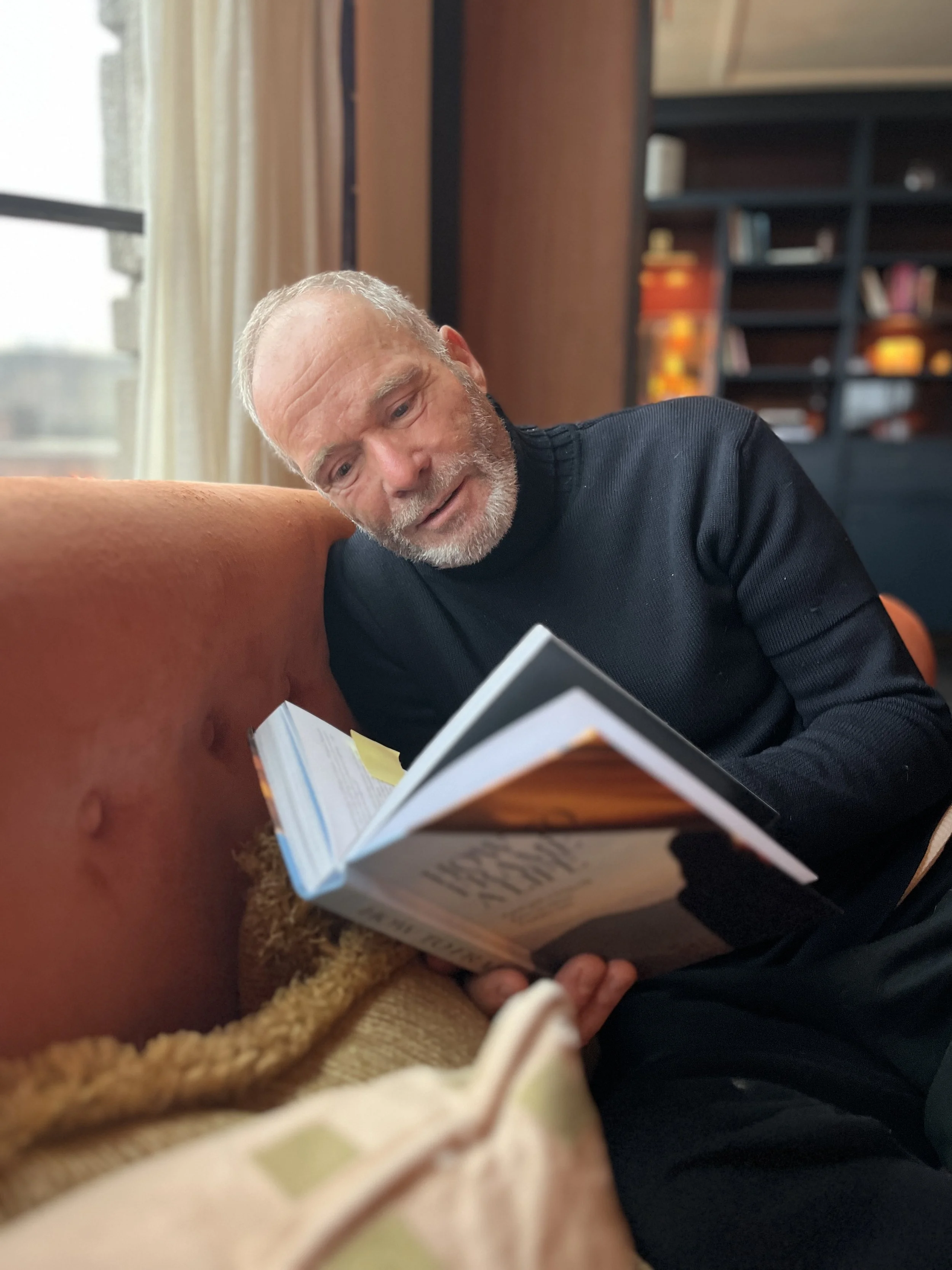 An elderly man with a gray beard and wearing a black sweater is sitting on a couch, reading a book, with a focused and engaged expression.