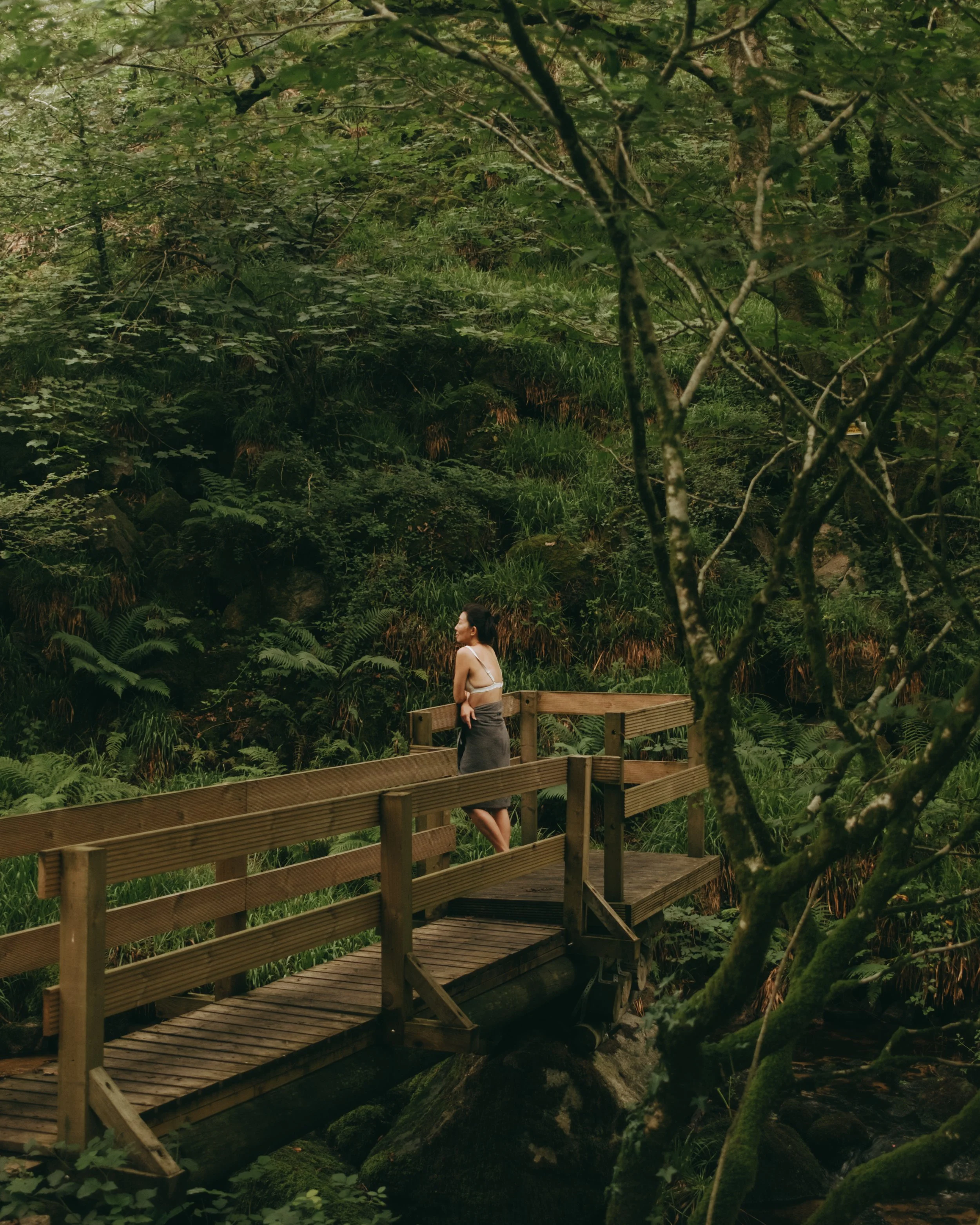 A woman standing on a wooden bridge in a lush green forest with dense foliage and trees.