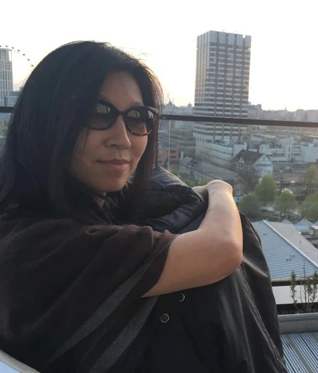 Woman with dark hair and sunglasses sitting on a rooftop with a city skyline in the background during sunset, hugging a black jacket.