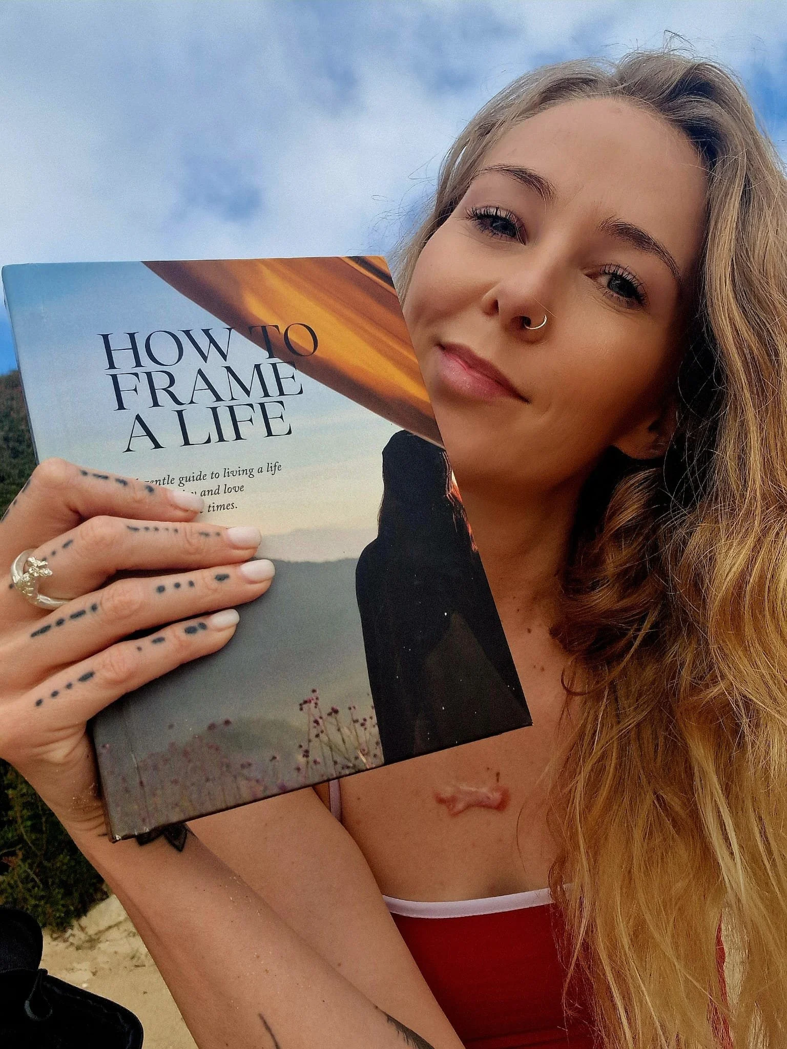 A young woman with light skin, long wavy hair, and a septum piercing holds a book titled "How to Frame a Life." She is outdoors against a partly cloudy sky, with a slight smile and tattoos on her hand and arm.