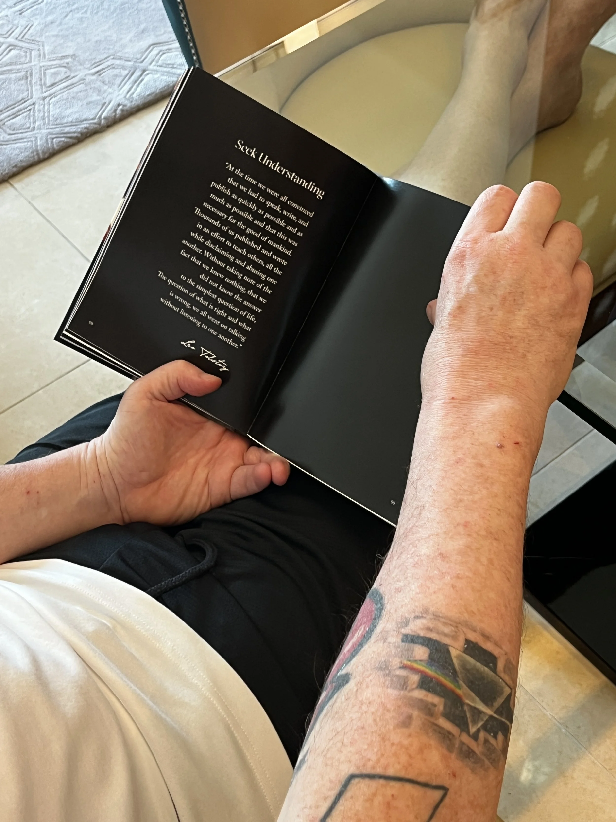 A person holding an open book with the title 'Sext Understanding.' The person has tattoos on their arm and is sitting at a glass table, reading the book.