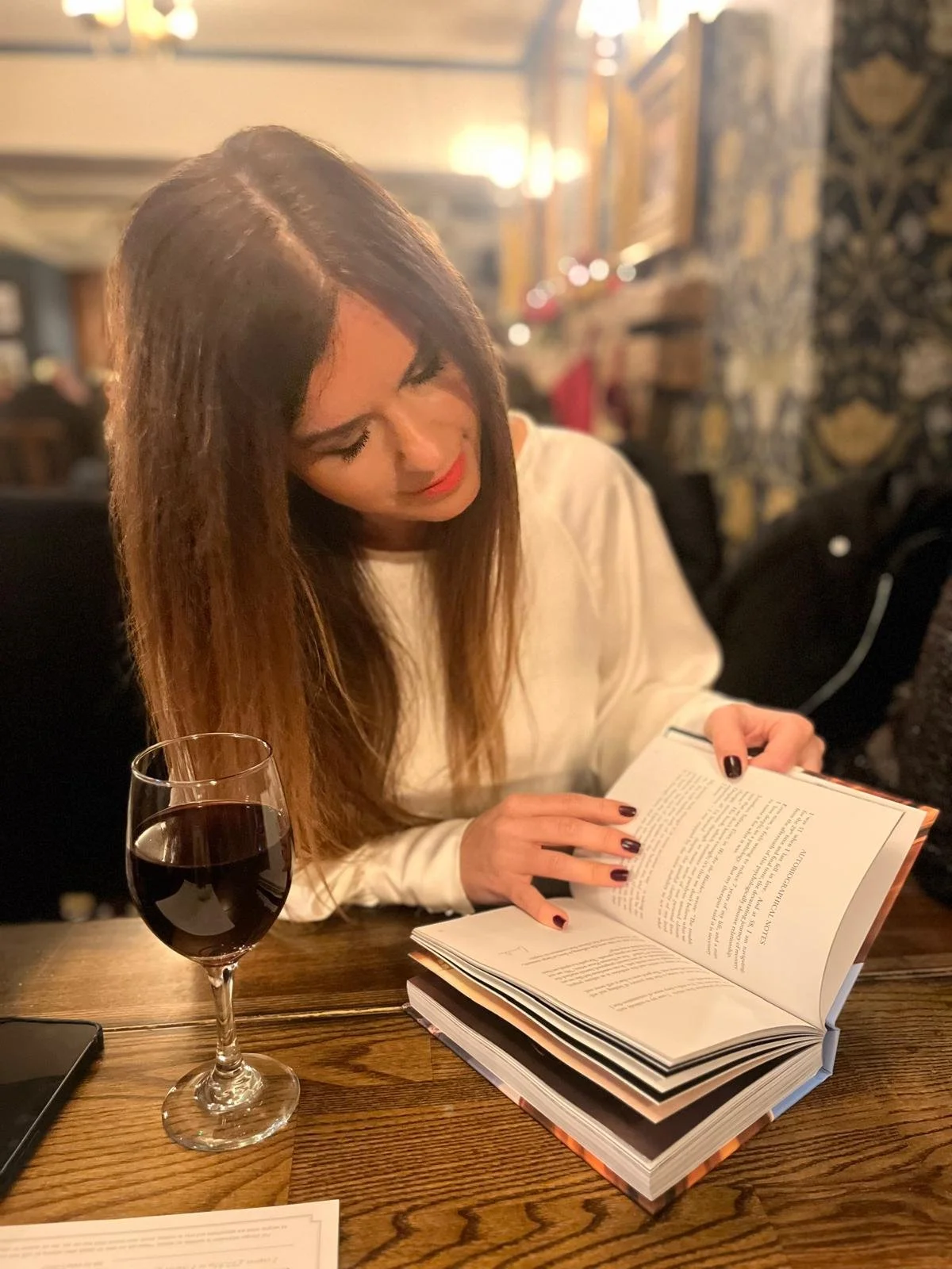 A woman with long brown hair and red lipstick reading a book at a restaurant table, with a glass of red wine in front of her, in a warmly lit setting.
