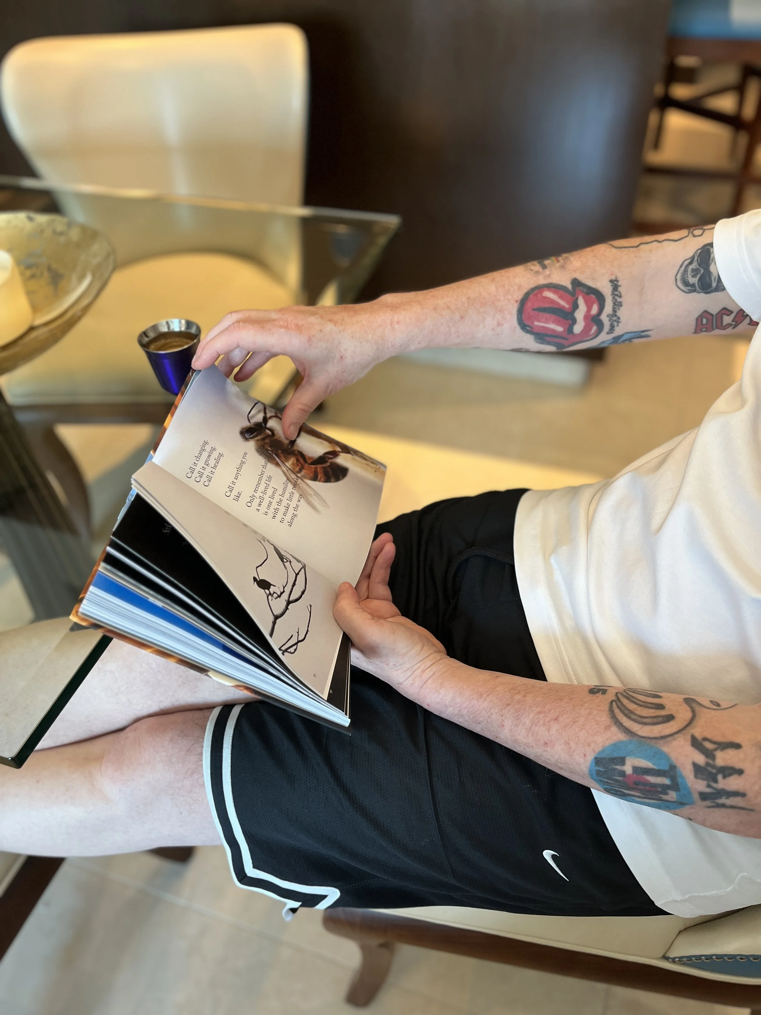 Person sitting at a table reading a book, wearing a white T-shirt and black shorts, with tattoos on their right arm. There is a glass and a bowl on the table.