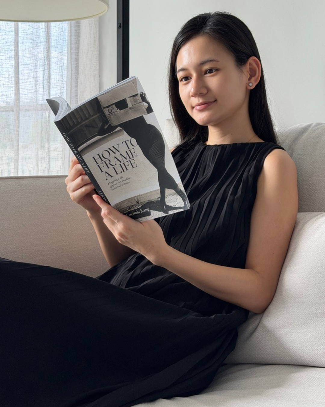 Young woman sitting on a beige sofa, reading a book titled 'How to Frame to a Life' with a black and white cover image