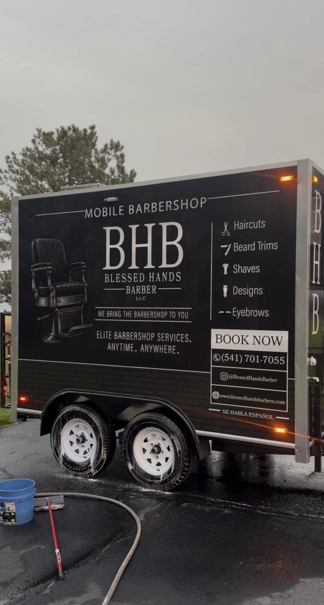 Black trailer advertising Blessed Hands Barber, a mobile barbershop business. The trailer features a barber chair graphic and text listing services like haircuts, beard trims, shaves, designs, and eyebrows. It includes contact information, social media handle, and website, with a button to book an appointment.