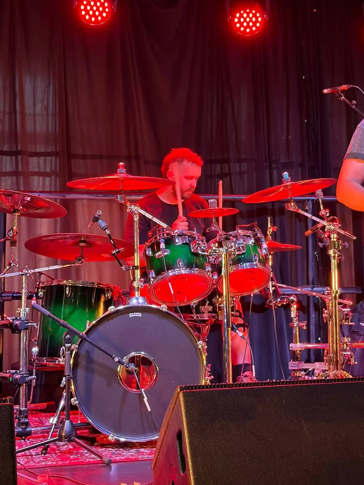 Male drummer playing a green drum set with red lighting on stage.