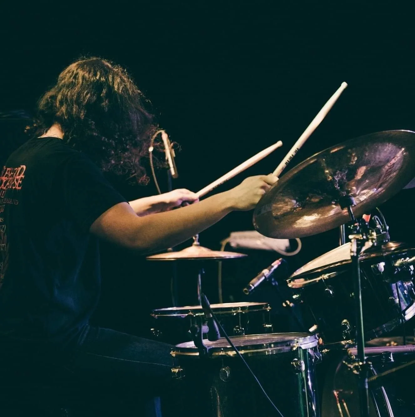 A musician with curly hair playing a drum set on stage, holding drumsticks, with a dark background.
