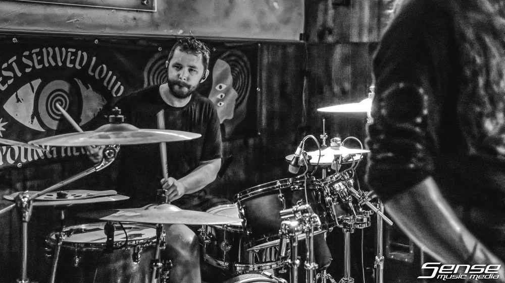 A drummer with a beard playing a drum set on stage during a live performance.