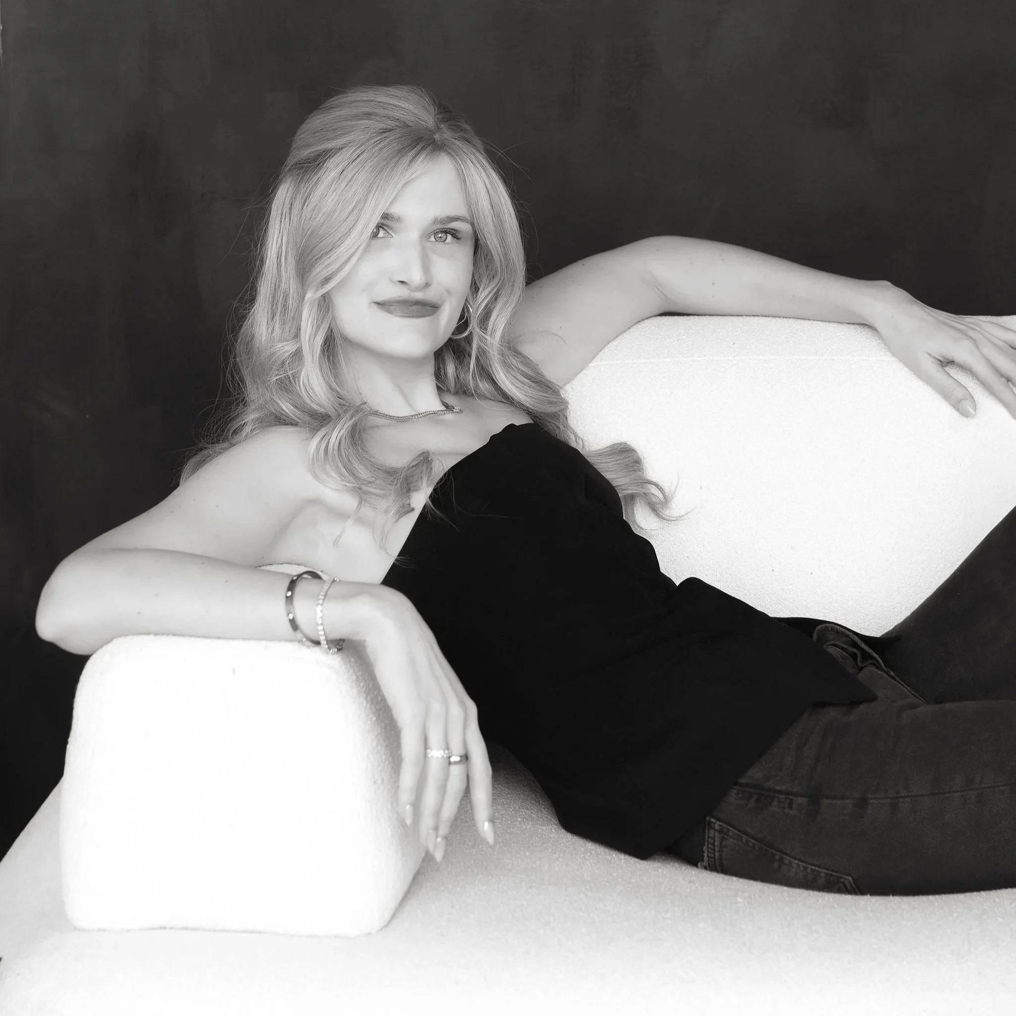 A woman with long, wavy hair reclining on a white chair, wearing a black strapless top, with her arm draped over the back of the chair, looking at the camera.