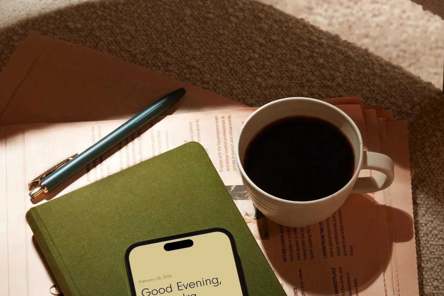 A study table with a blue pen, a green notebook, a smartphone displaying "Good Evening," a cup of black coffee, and some printed papers on a beige carpeted surface.