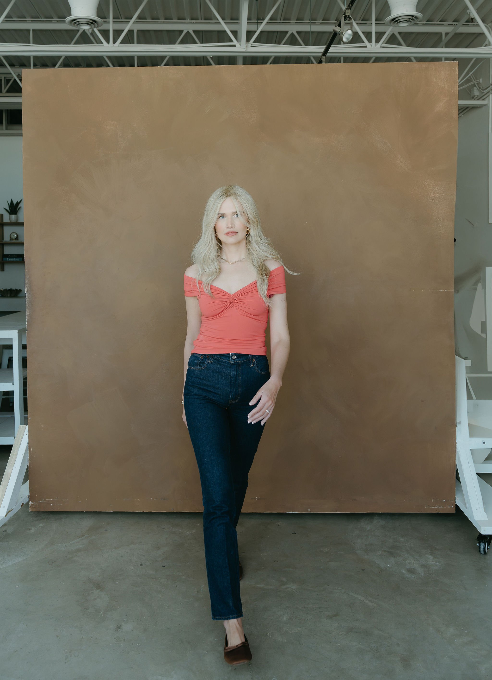 A woman with long blonde hair, wearing a red off-shoulder top and dark blue jeans, standing in front of a brown painted background in an indoor studio.