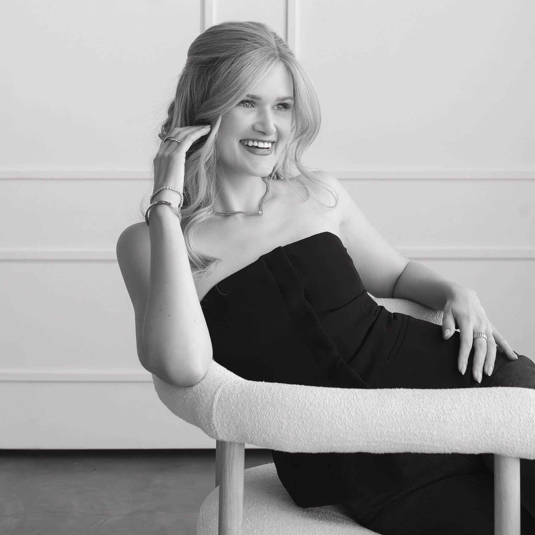 A woman wearing a strapless black dress sitting in a modern chair, smiling, with her hand near her face, in a bright room with minimal decor.