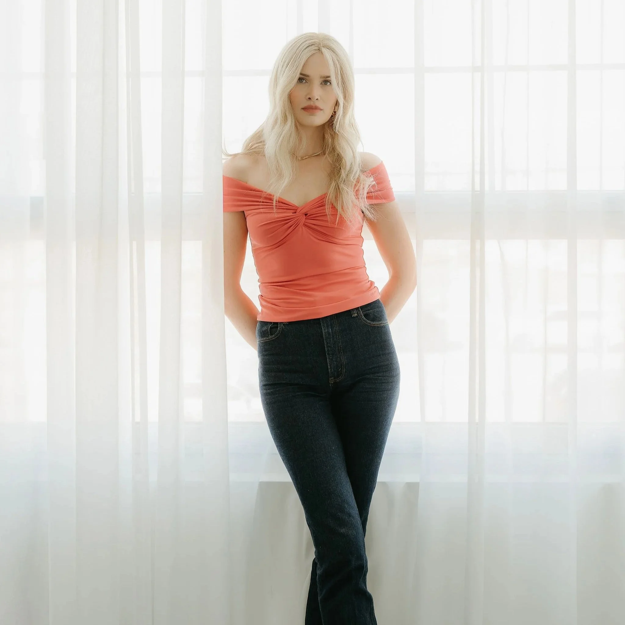Young woman with blonde hair wearing a pink off-shoulder top and dark jeans standing in front of sheer white curtains.