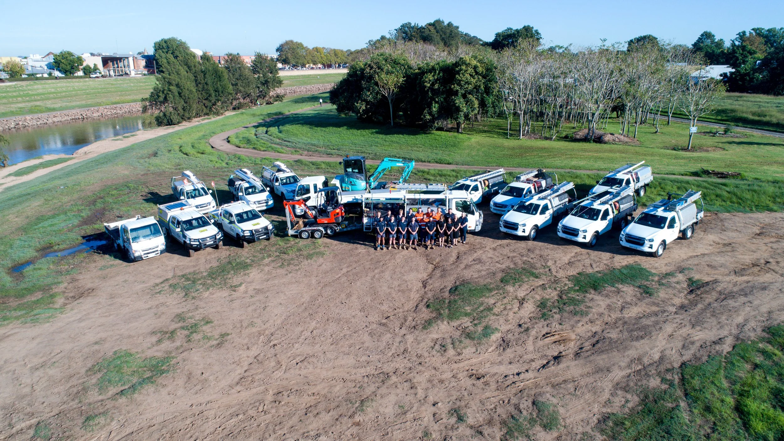 A group of workers standing in front of work trucks and construction equipment in an open field with trees and a river in the background.