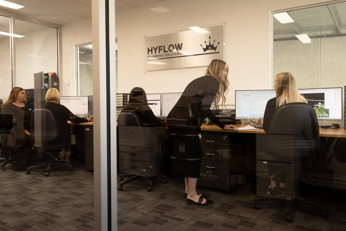 People working in an office at computer desks, seen through a glass wall, with a woman standing and talking to a seated woman.