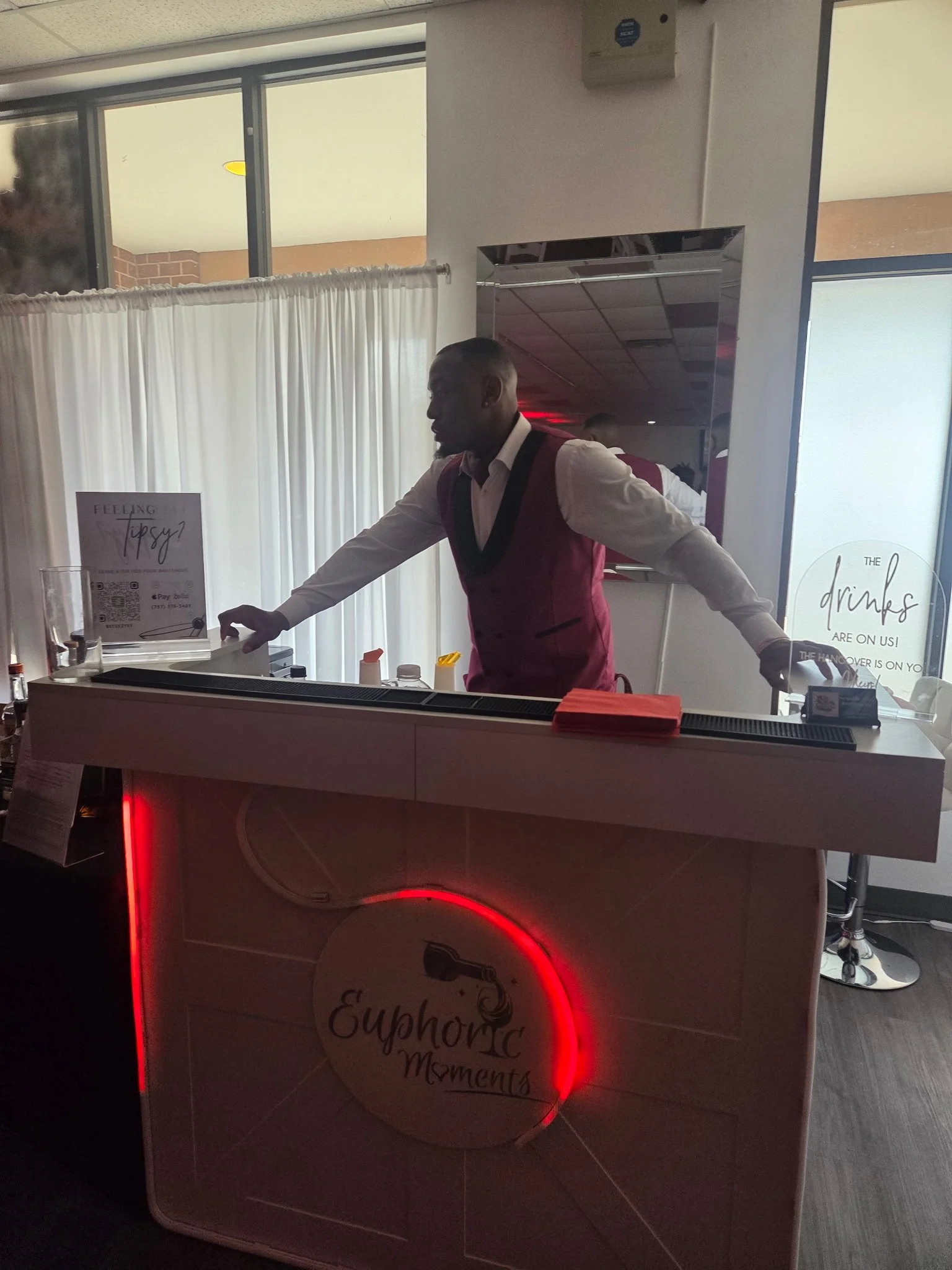 A man in a white shirt and red vest behind a bar at a social event, with signage that reads 'Euphoria Moments' and a poster mentioning 'tipsy' and 'drinks are on us' in the background.