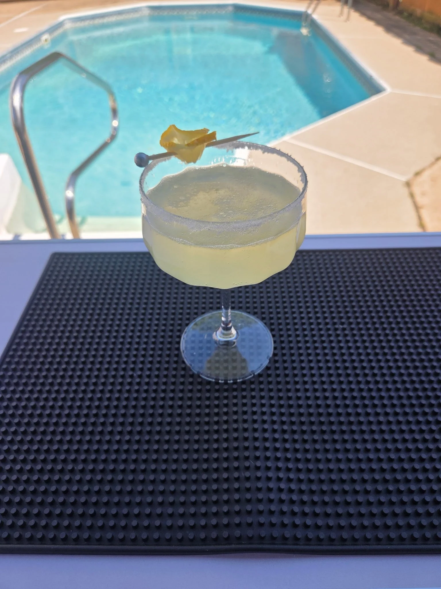 A cocktail glass with a salted or sugar rim and a slice of pineapple or lemon on the edge, placed on a black textured surface such as a bar mat by an outdoor swimming pool.