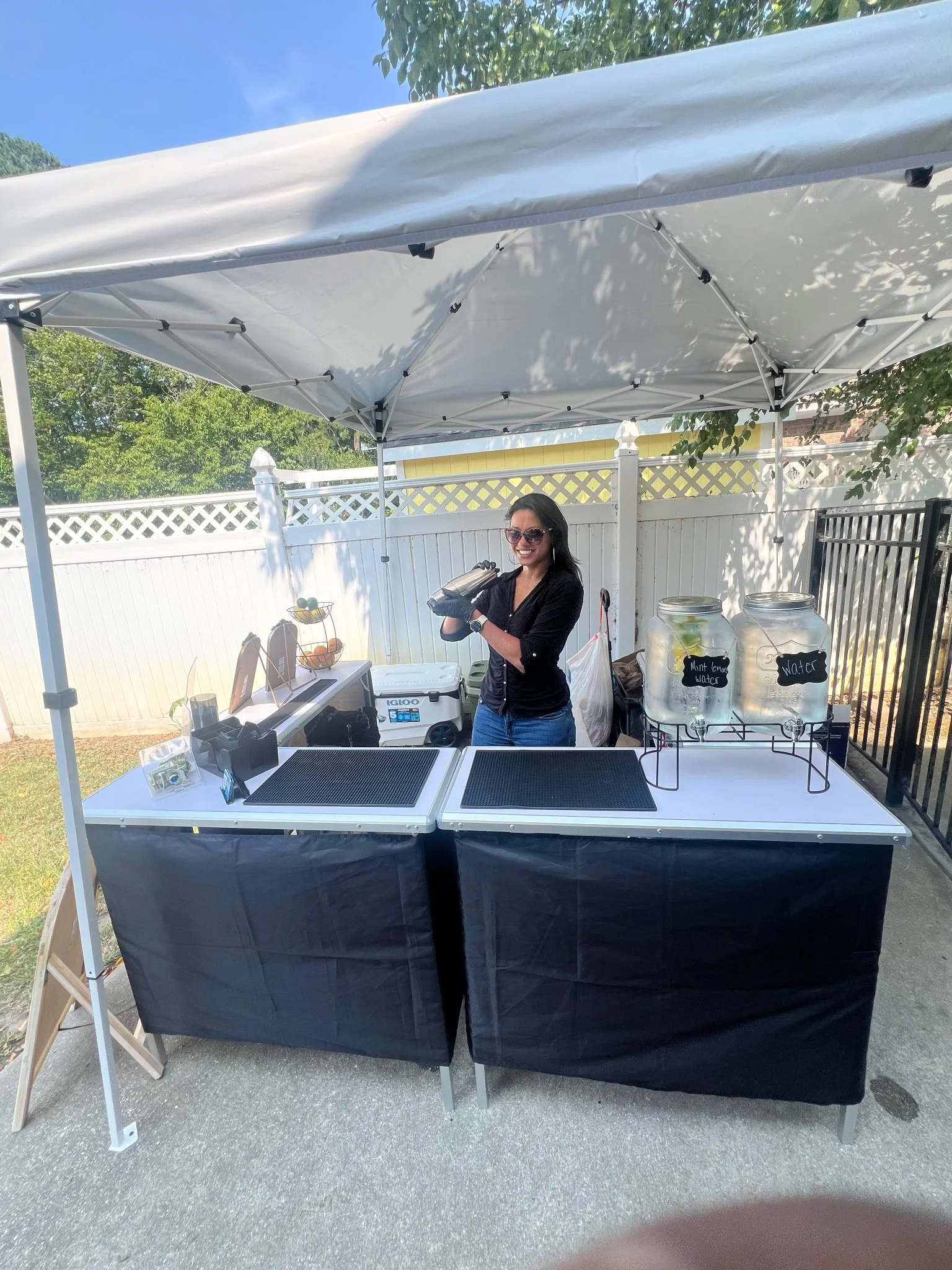A woman standing behind a table at an outdoor mobile bar station, wearing sunglasses and smiling, under a white canopy tent with likely liquors, garnishes, mixers, etc. underneath. The table has jars labeled 'Mint Lemon Water' and 'Water' with jars o