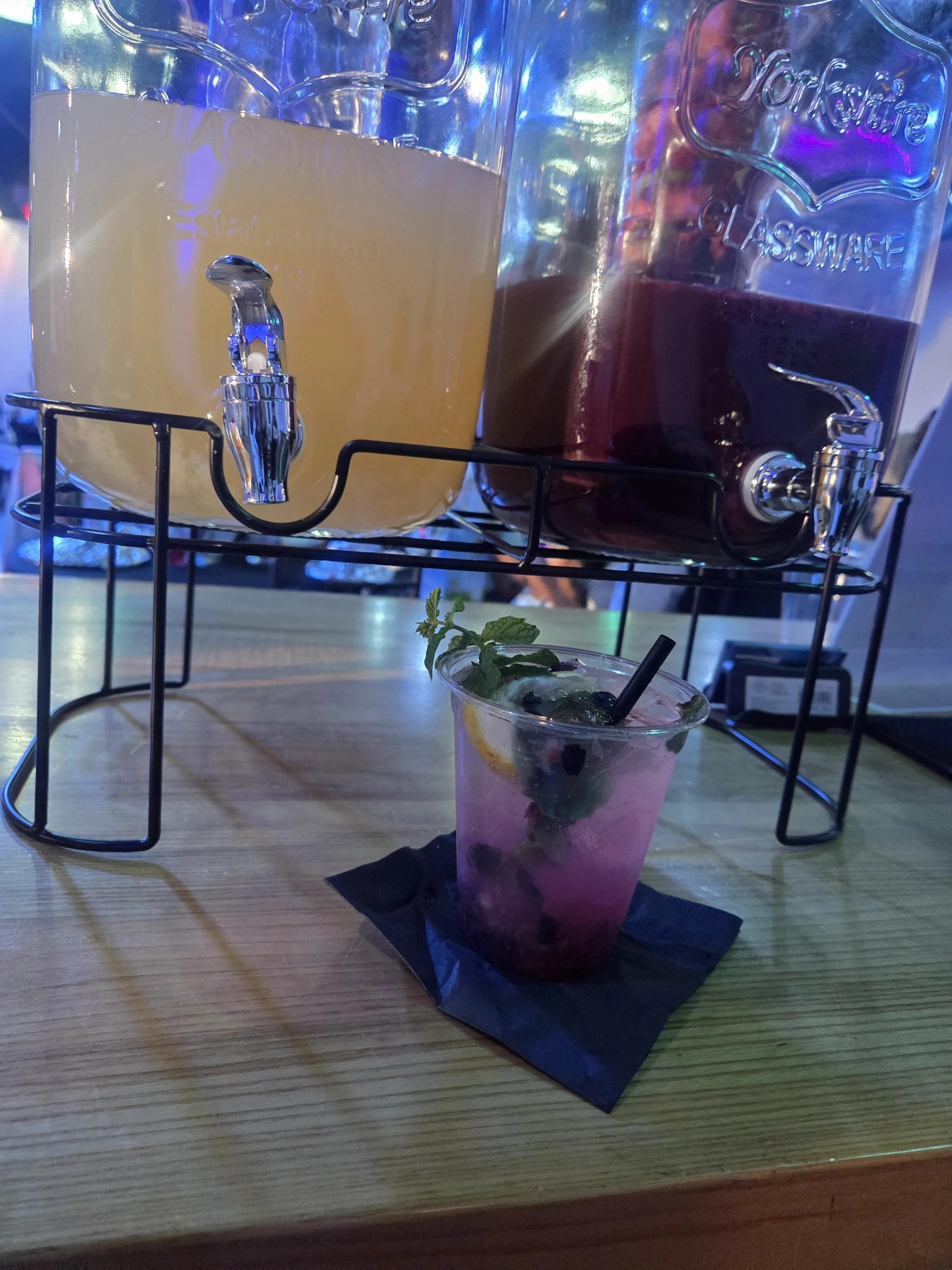 A glass pitcher with a spigot filled with margarita, and a glass pitcher filled with sangria on a metal stand. In the foreground, there is a pink cocktail with blueberries, mint, lemon, and a black straw, placed on a black napkin on a wooden table in