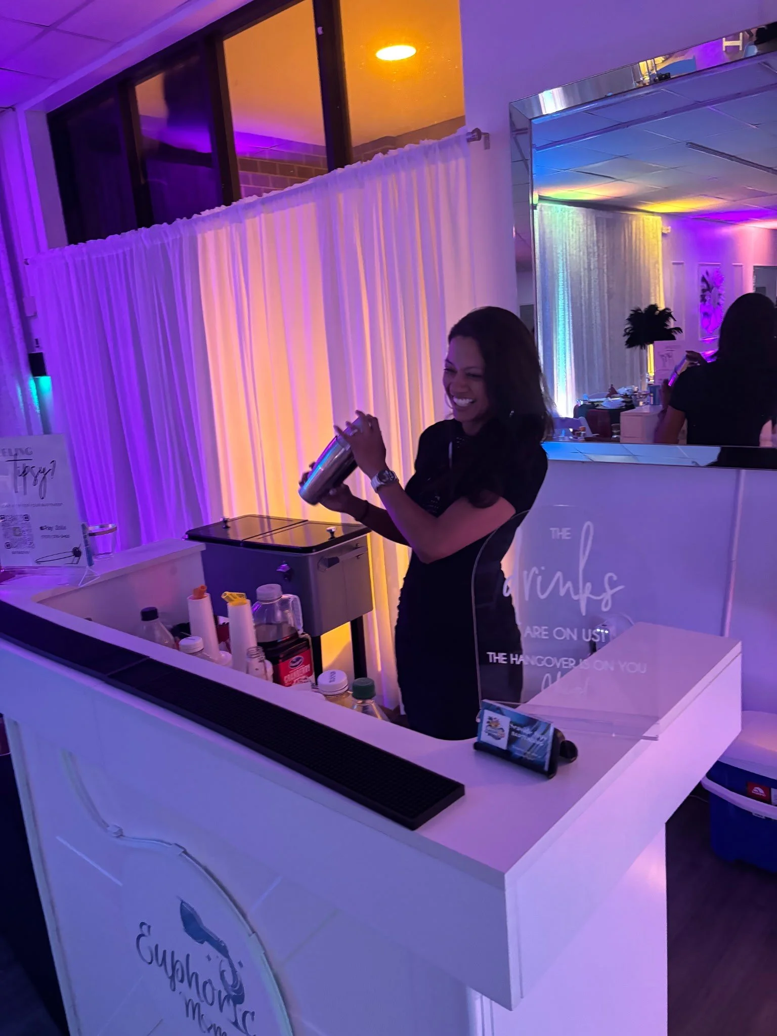 A woman smiling and holding a cocktail Boston shaker at an event with colorful LED lighting, a white bar counter with various bottles and supplies, and draped curtains in the background.