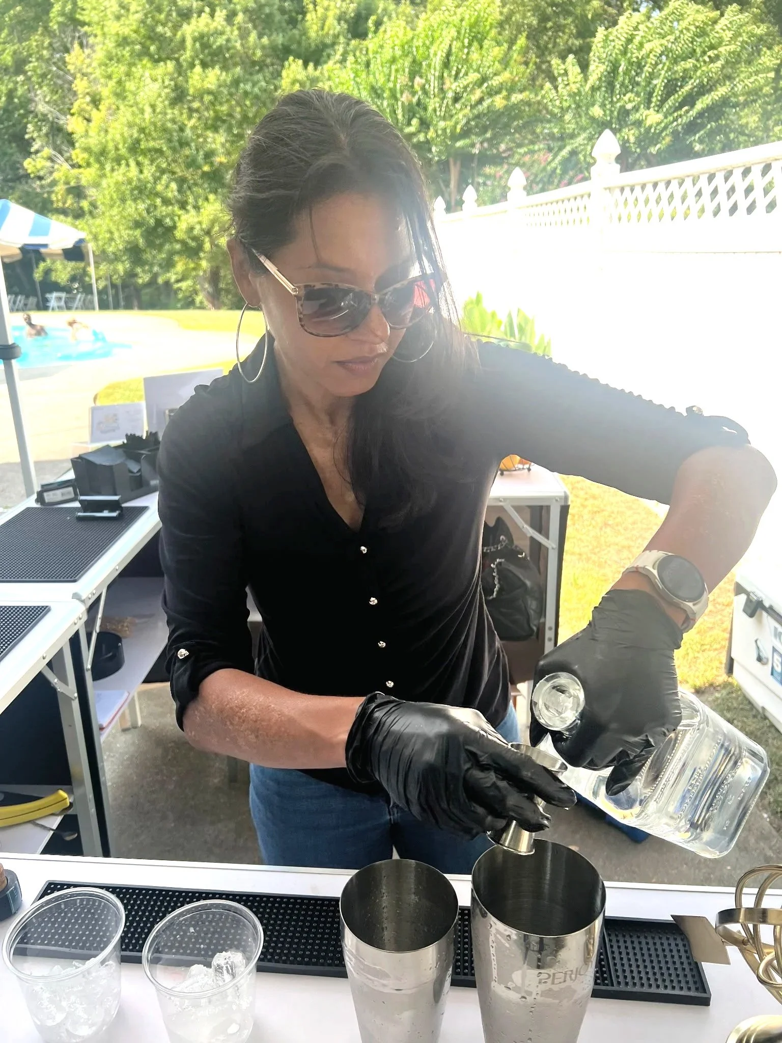 A woman with long dark hair, wearing sunglasses, hoop earrings, a black button-up top, and a watch, is seen pouring liquid into a container. She is working outdoors with a white fence and green trees in the background. There are various bar tools and cups on the bar counter in front of her.