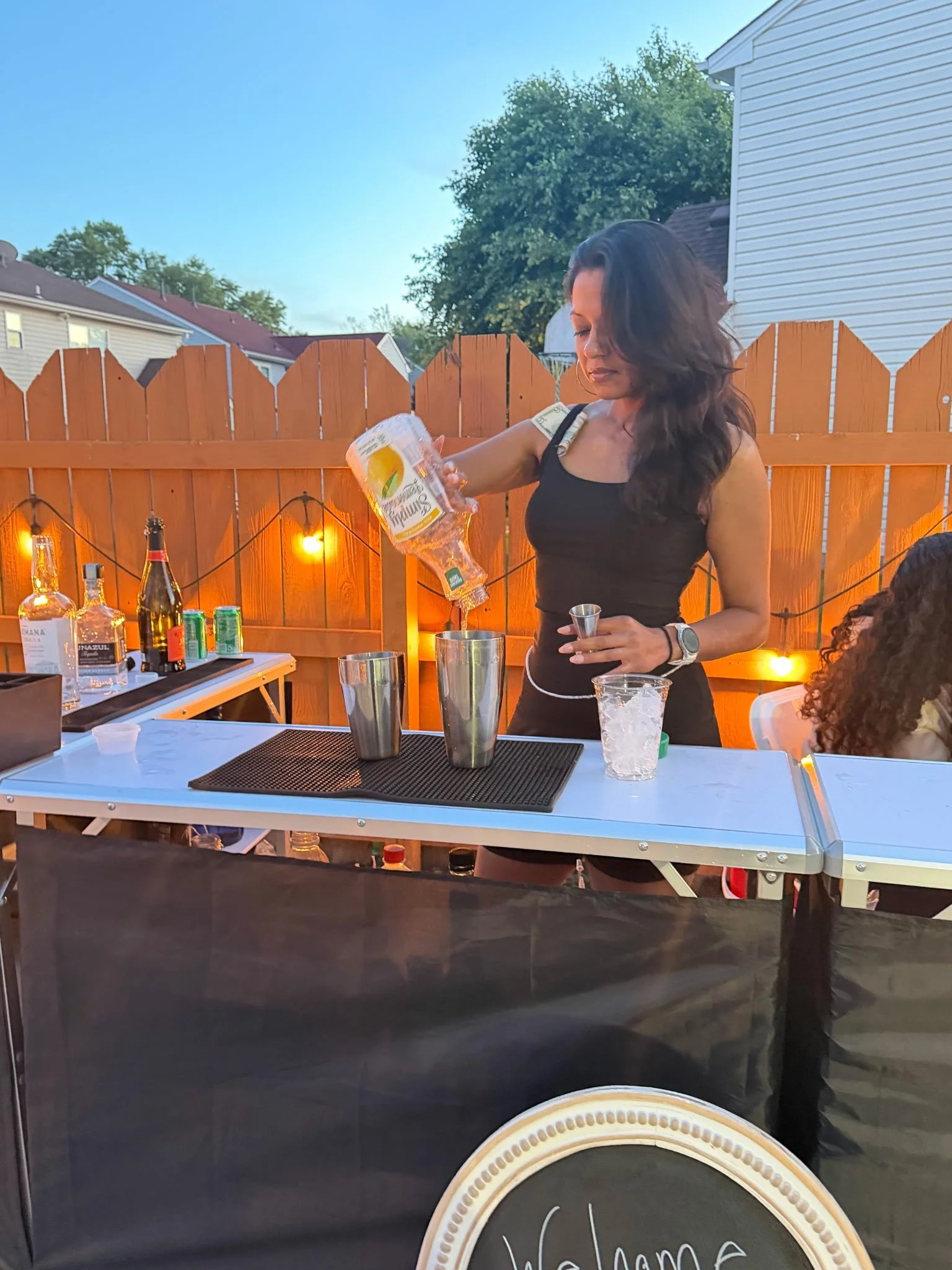 A woman with long dark hair wearing a black tank top and hoop earrings pours a drink into a clear plastic cup at an outdoor gathering during the evening. The setting features a wooden fence decorated with string lights, and a white table with various bottles and bar tools. Part of a decorative sign reading 'Welcome' is visible at the bottom.